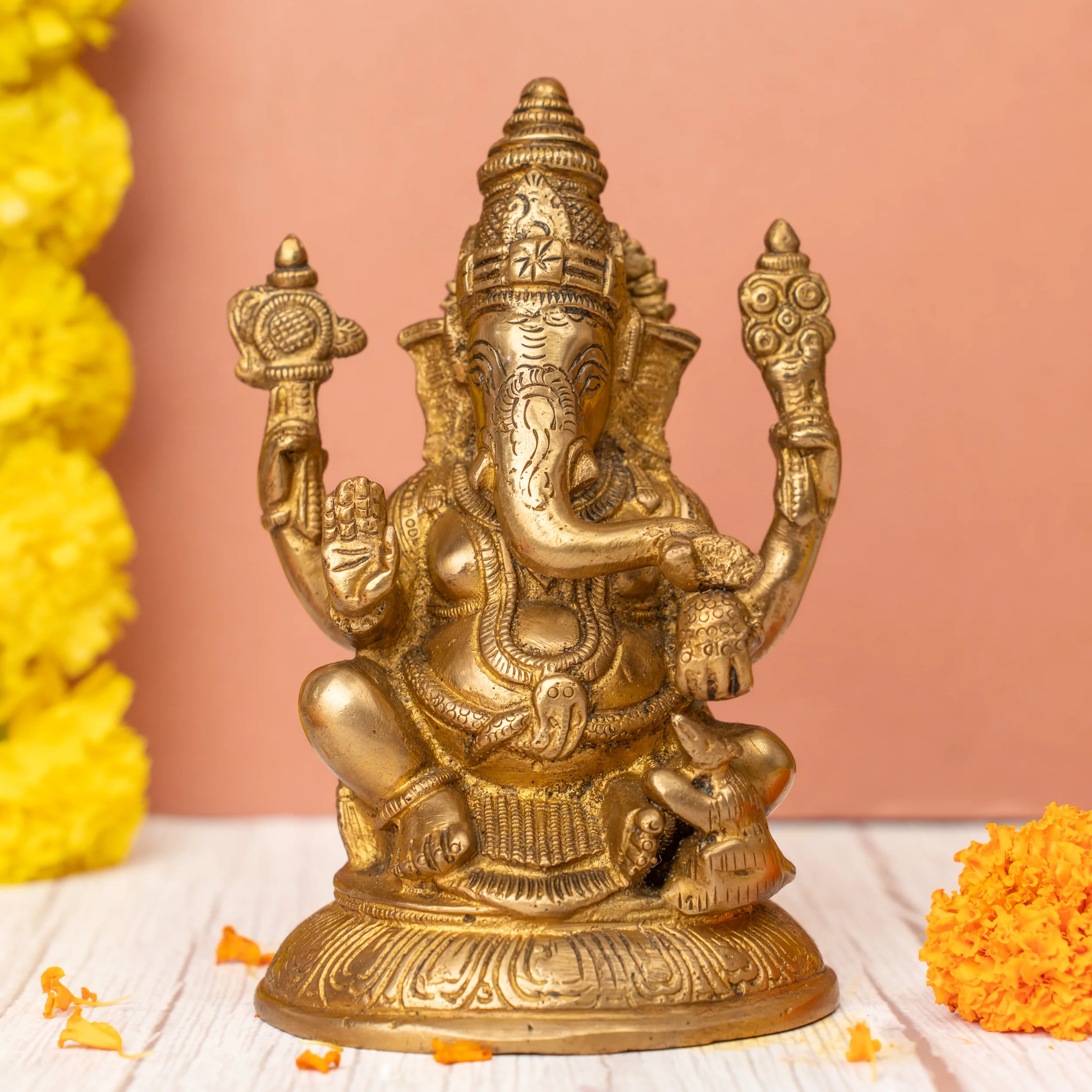 Golden Ganesha idol with multiple arms on a white surface, orange and yellow flowers beside it, pink background.
