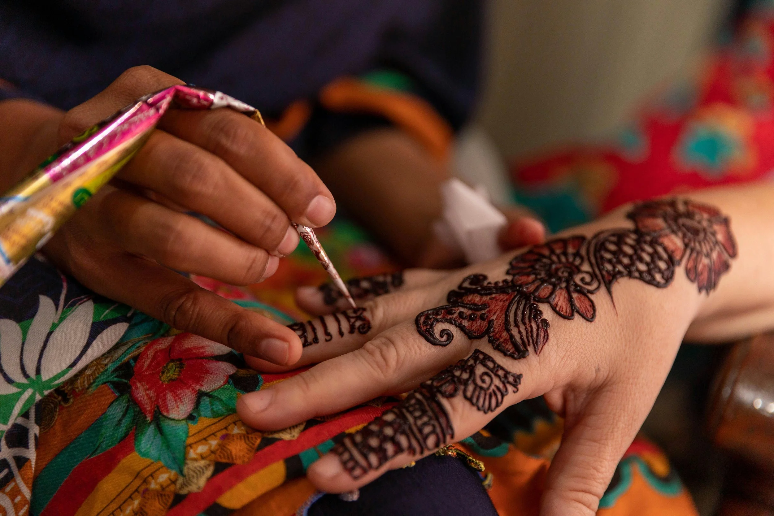 A person getting a henna tattoo on their hand, with the artist applying the design using a thin cone and black henna, while wearing colorful clothing with floral patterns.