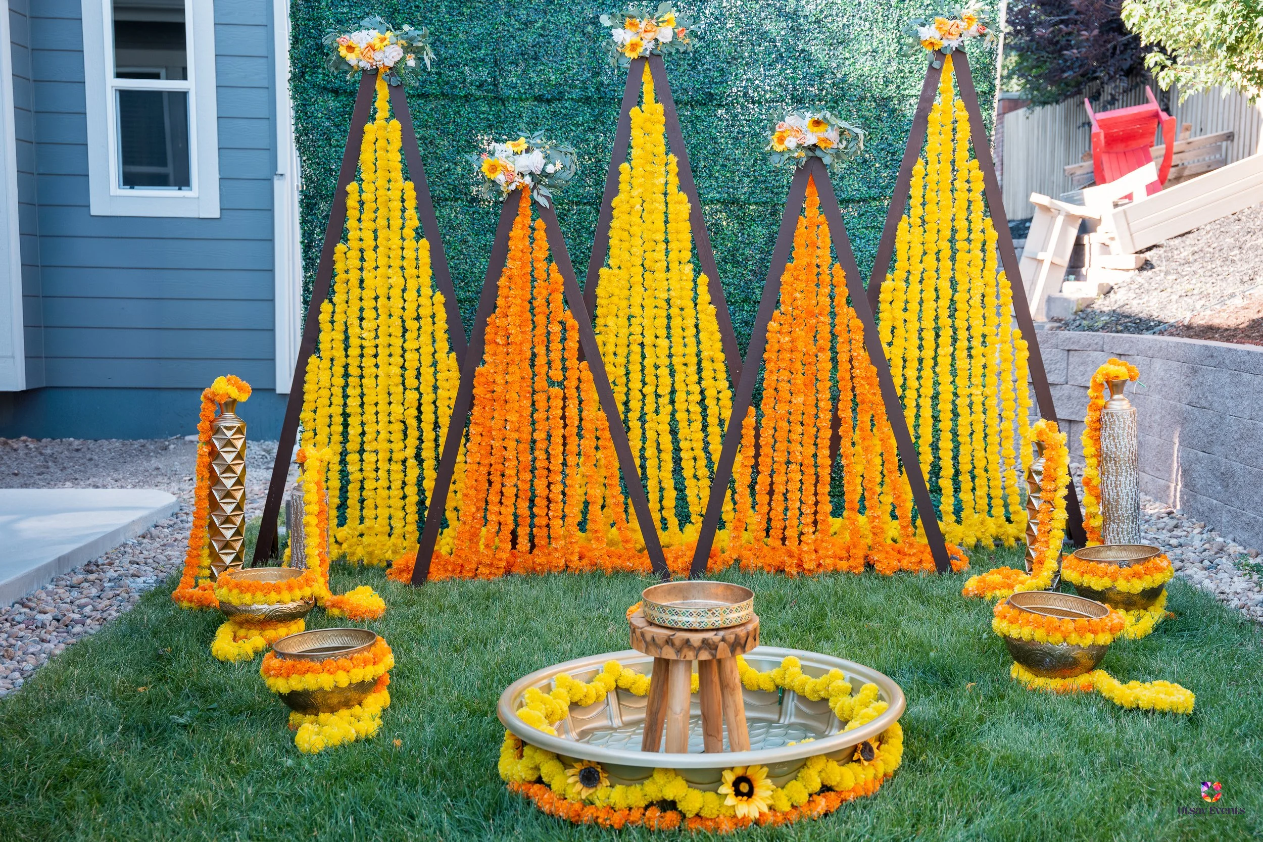 Decorative display of orange and yellow marigold flower garlands arranged in triangular patterns on wooden stands, with vases and bowls decorated with flower garlands on the grass in front, for a festive celebration outdoors.
