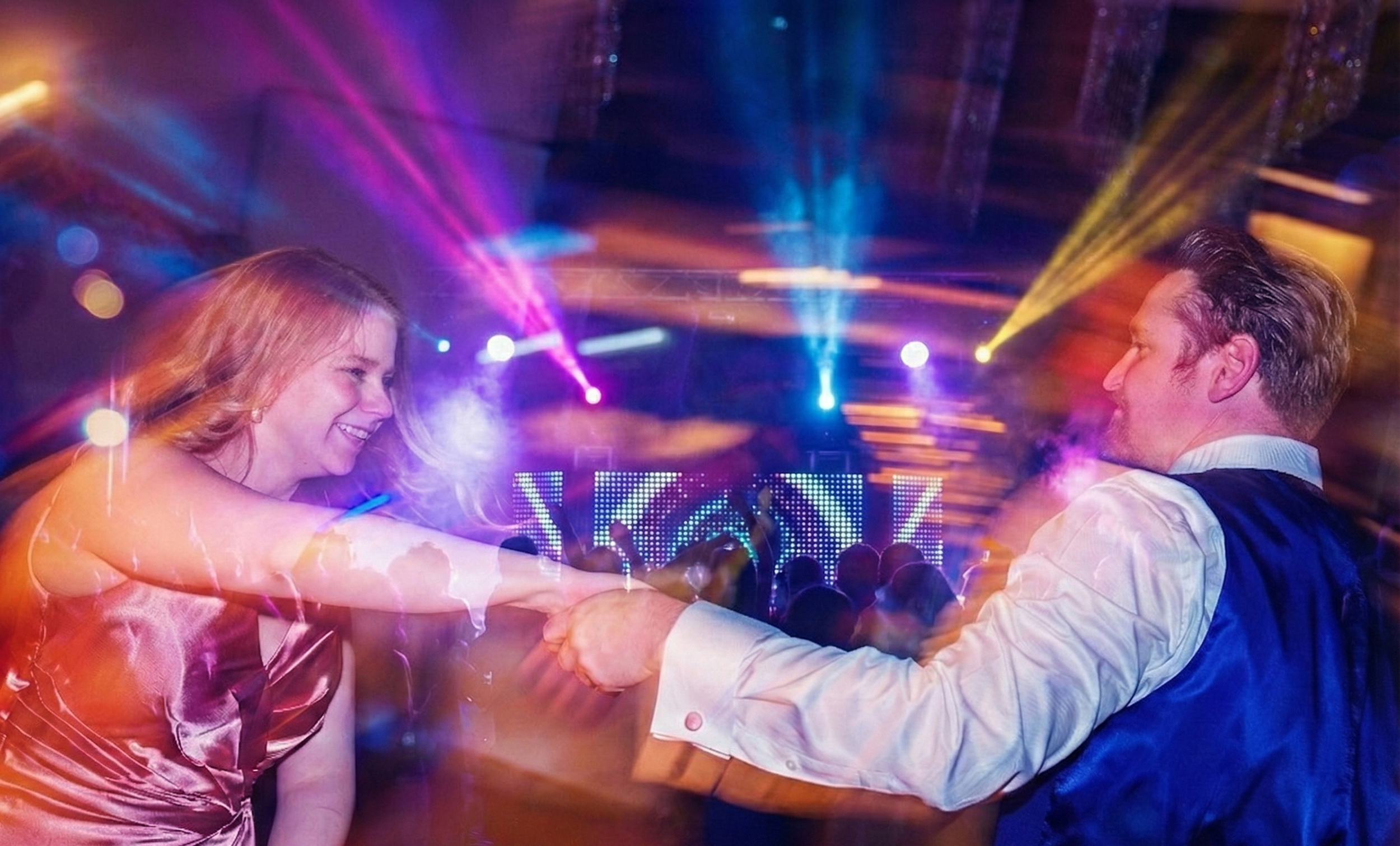 A man and woman dancing and smiling at a party with colorful lights and a DJ stage in the background.