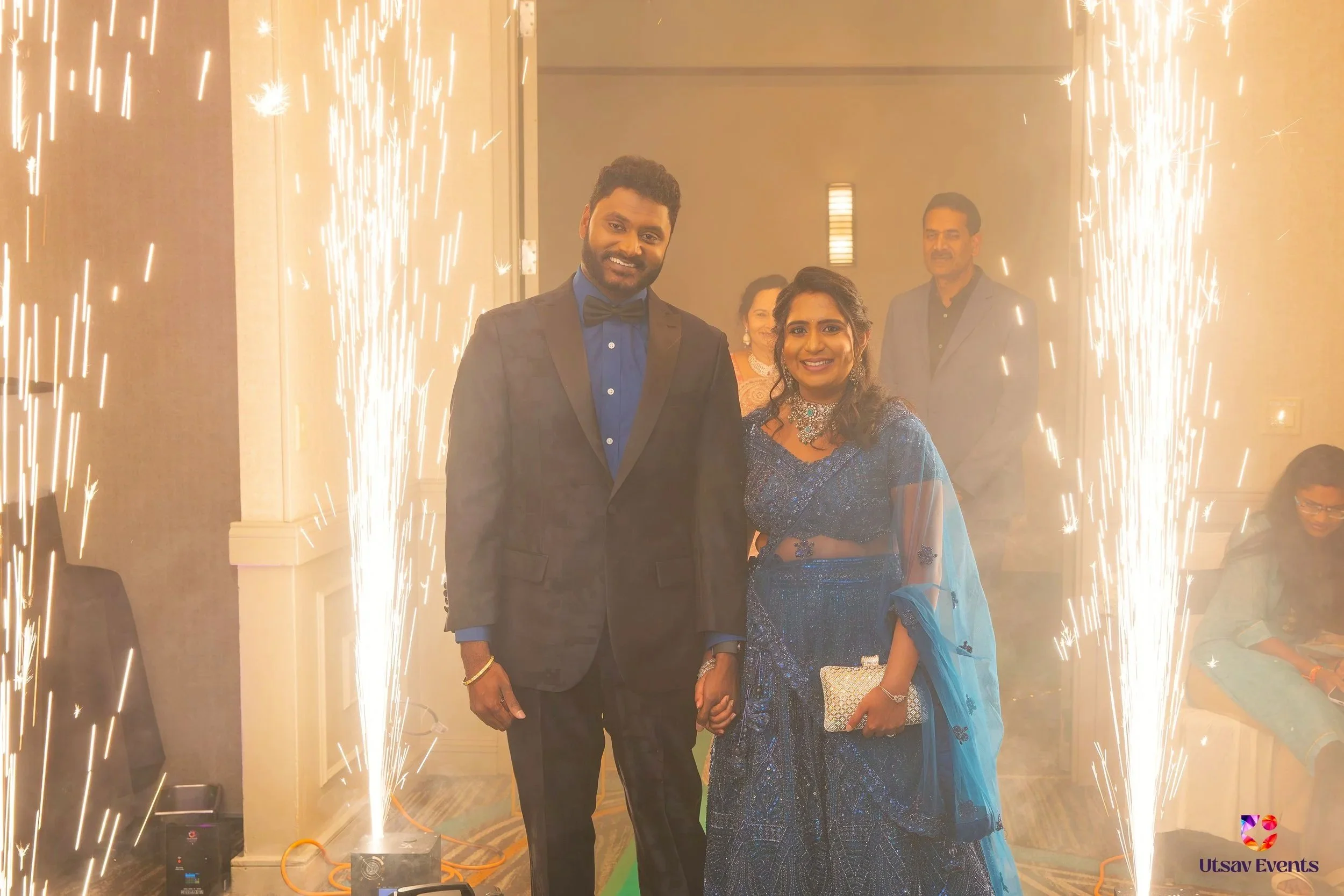 A smiling couple dressed in formal attire holding hands at a celebration, with sparklers and guests in the background.