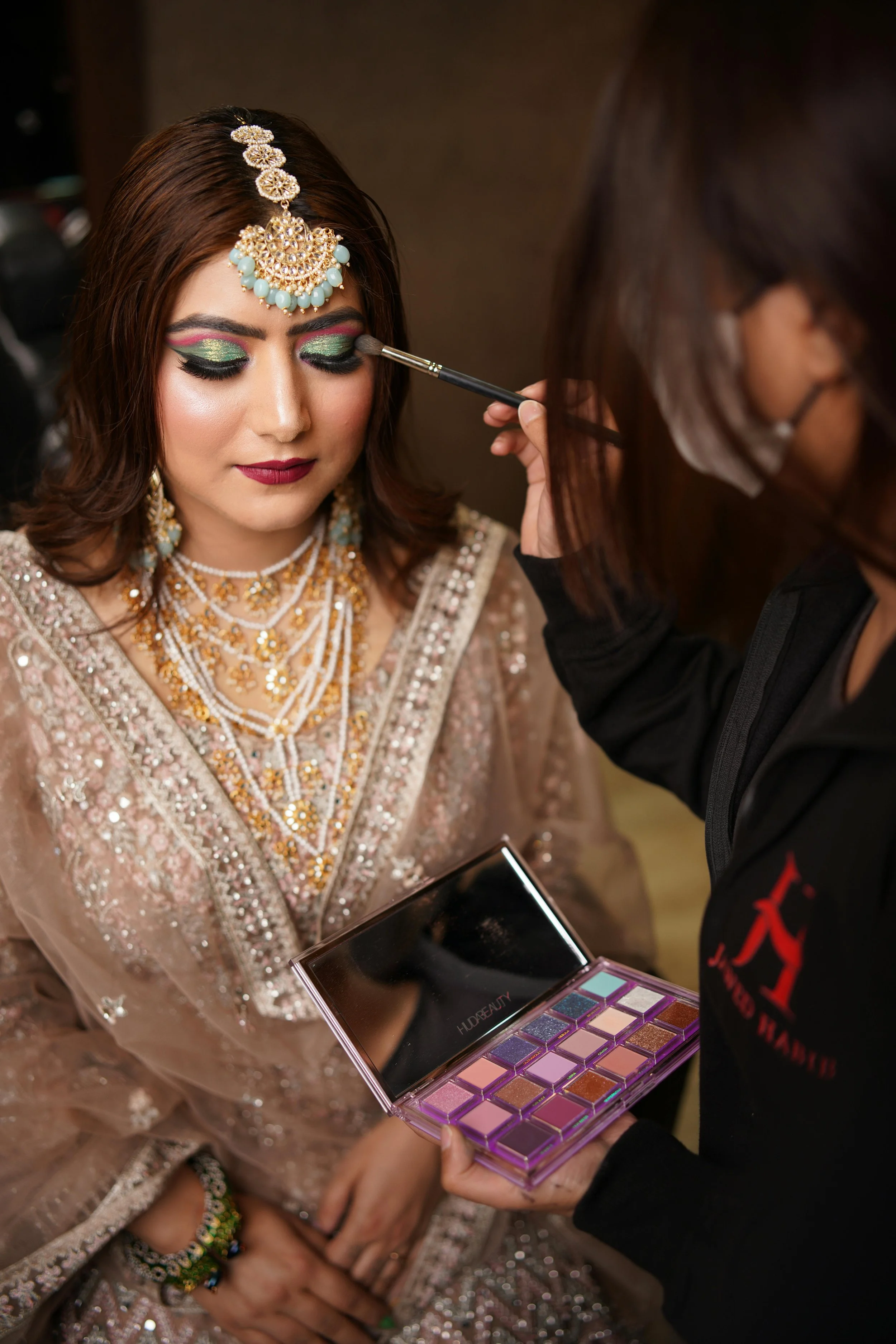 A woman in traditional Indian attire and jewelry is having her eye makeup applied by a makeup artist using eyeshadow from a palette.