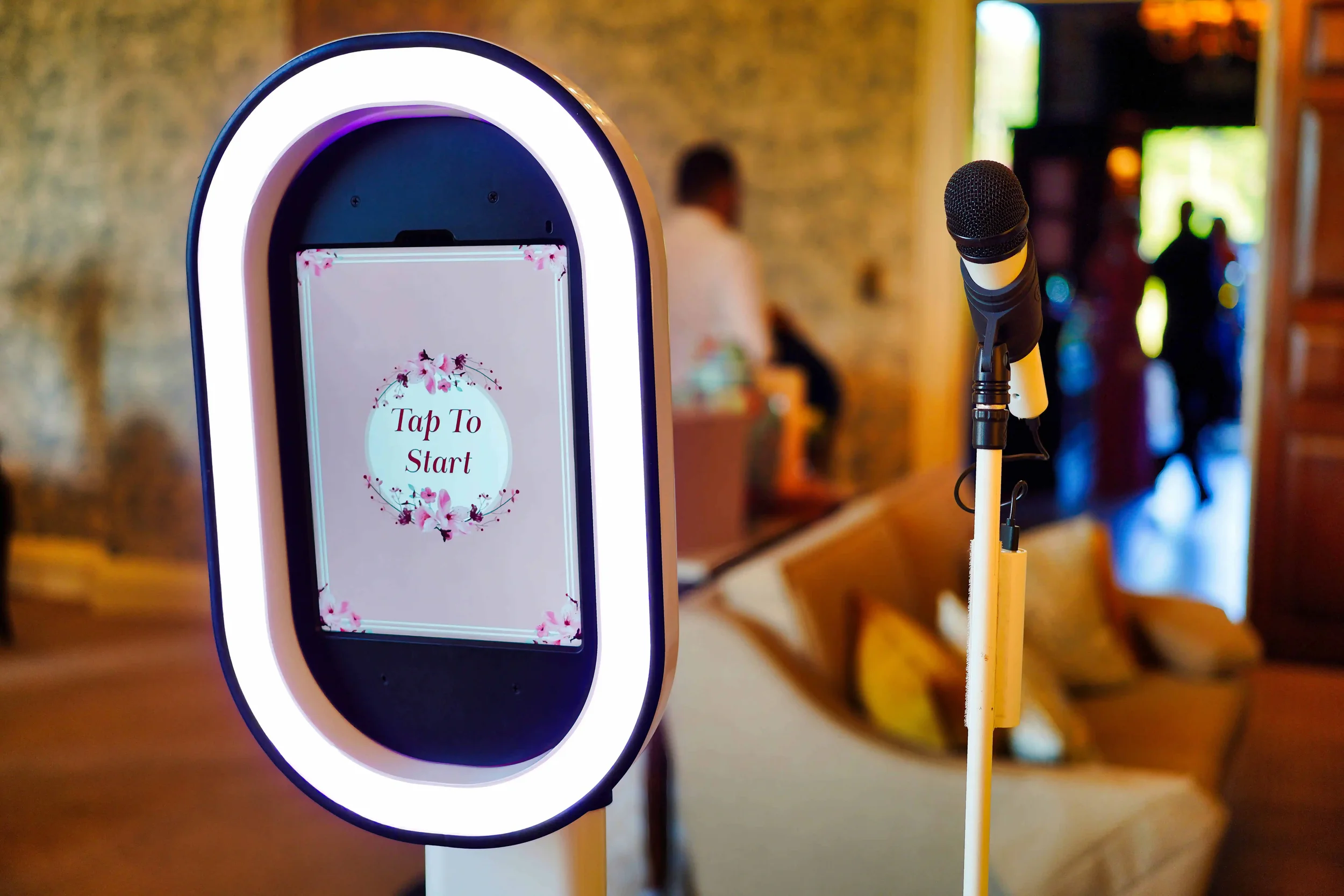 A digital photo booth with a circular screen displaying 'Tap To Start' decorated with pink floral designs. A microphone is positioned nearby, and the background shows a blurred indoor setting with people and a cozy seating area.