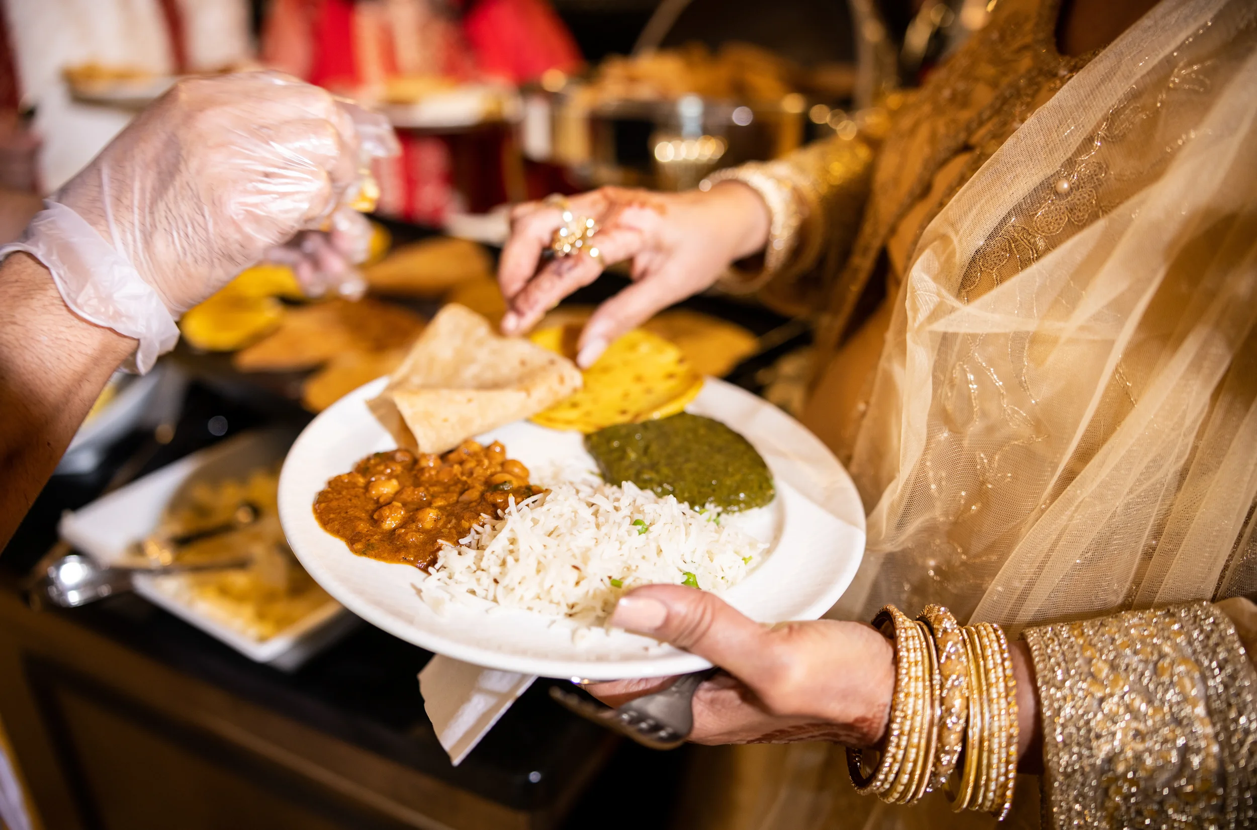 Close-up of a woman in traditional Indian attire holding a plate of Indian food, with another person serving food in the background.