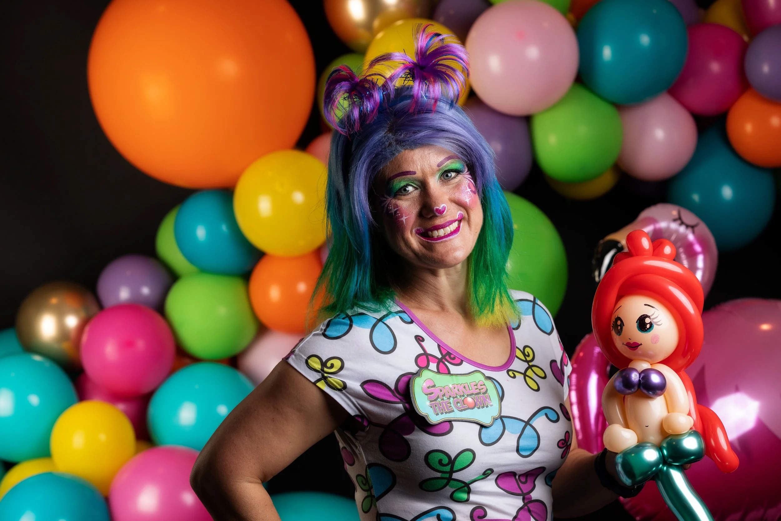 A woman with colorful rainbow hair, wearing vibrant makeup and a fun shirt with a star badge, stands in front of a backdrop of multicolored balloons while holding a balloon art figure of a girl with red hair and a purple top.
