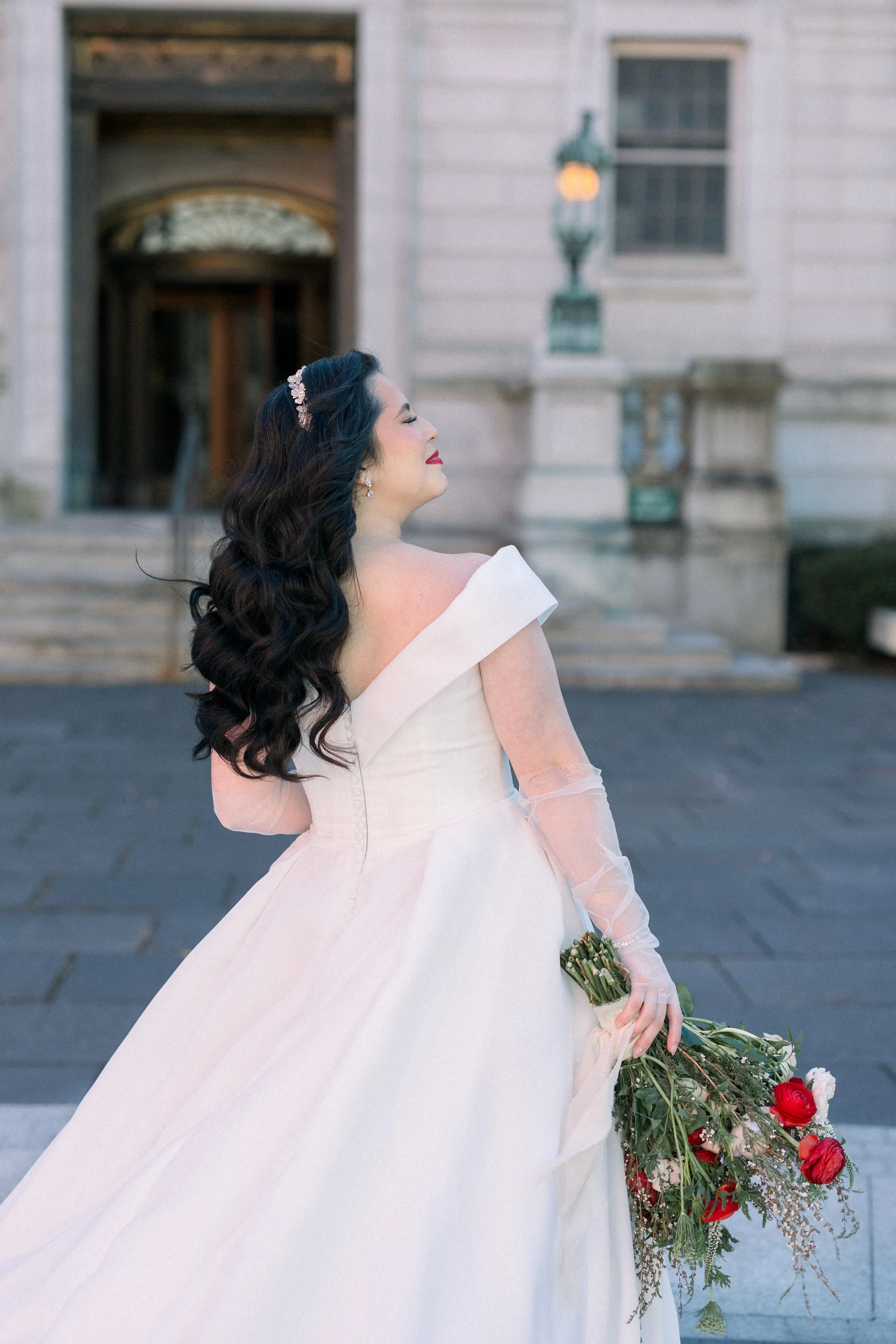 Bride in a white wedding dress with off-the-shoulder neckline holding a bouquet of red and white flowers, standing outside a building with stairs and a lamp post, with her eyes closed and a serene expression.