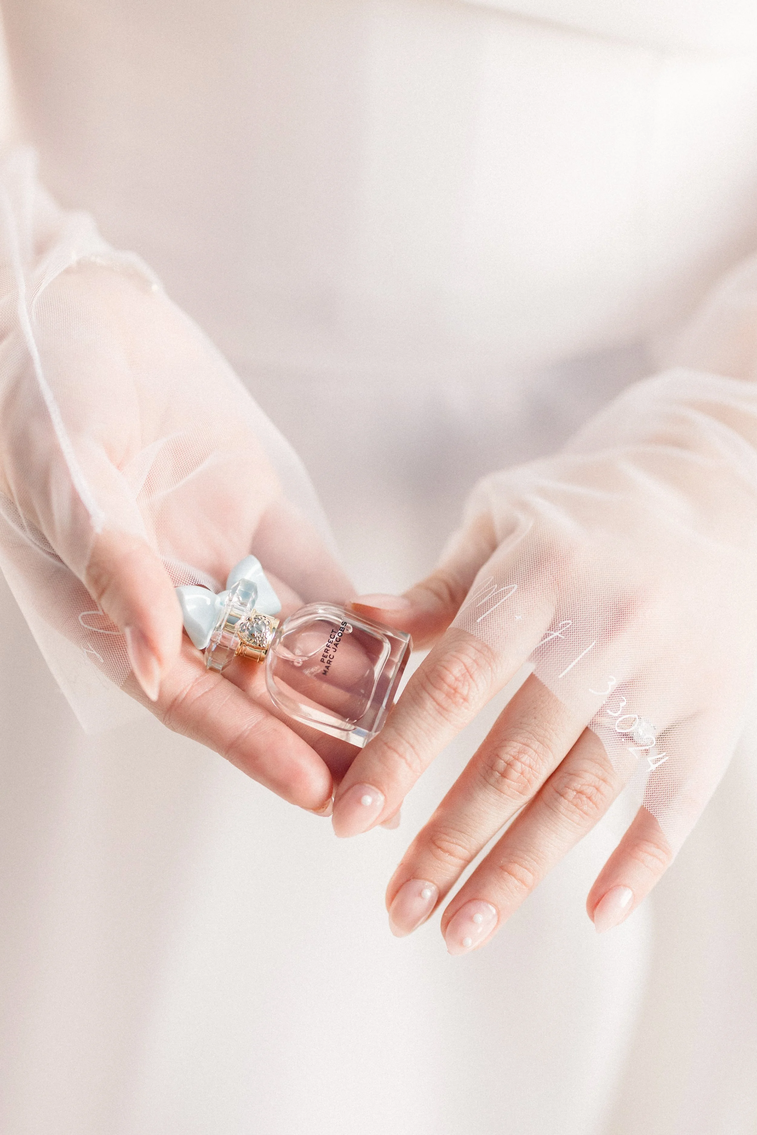 A person wearing sheer, light-colored fabric holding a small, decorative perfume bottle with a bow on the cap.