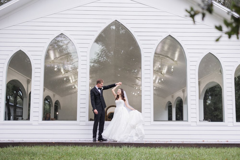 Groom twirls his bride in front of the white chapel with arched windows at Whitewood Ranch wedding venue