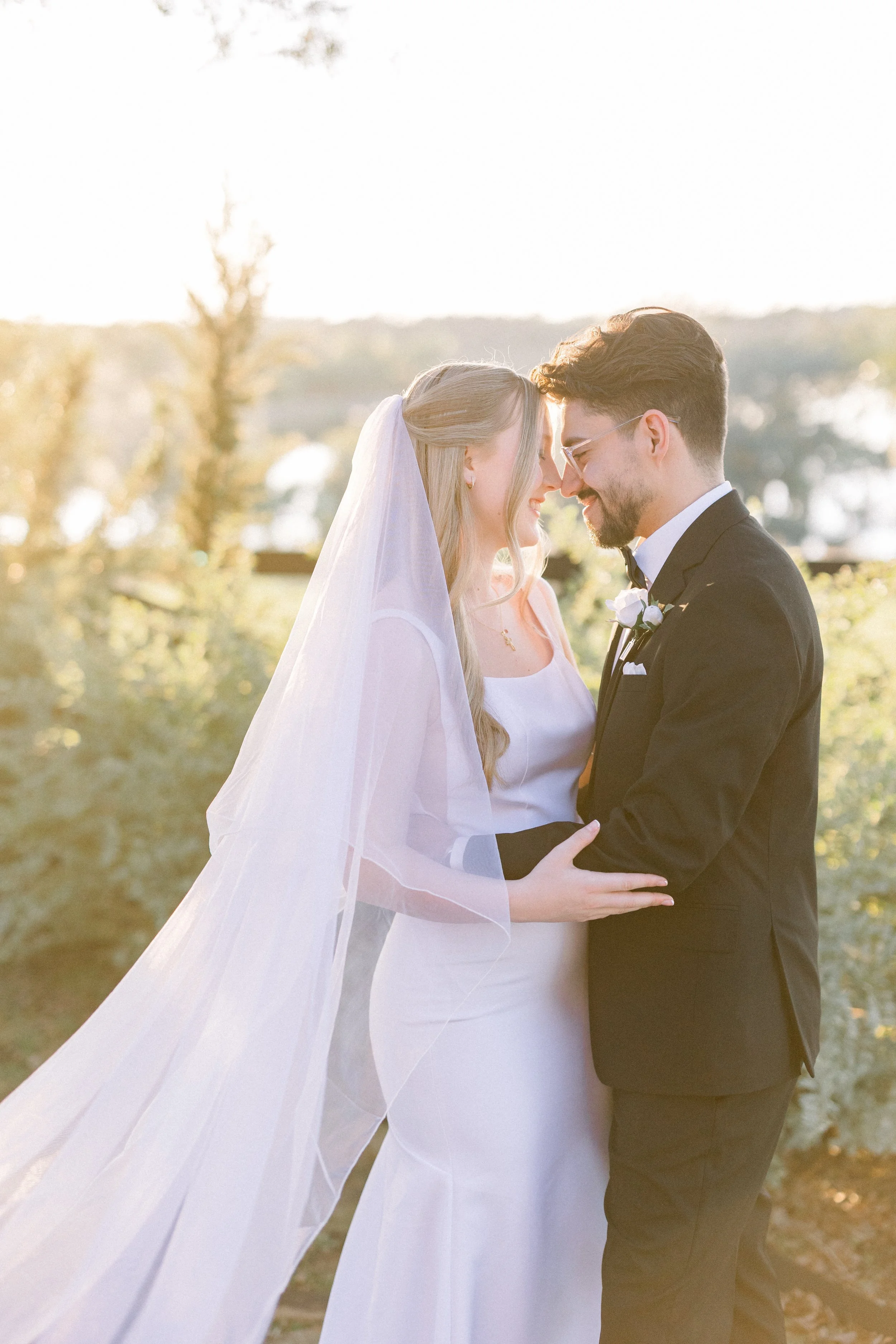 A newlywed couple dressed in wedding attire sharing a tender moment outdoors during golden hour, with trees and soft sunlight in the background.
