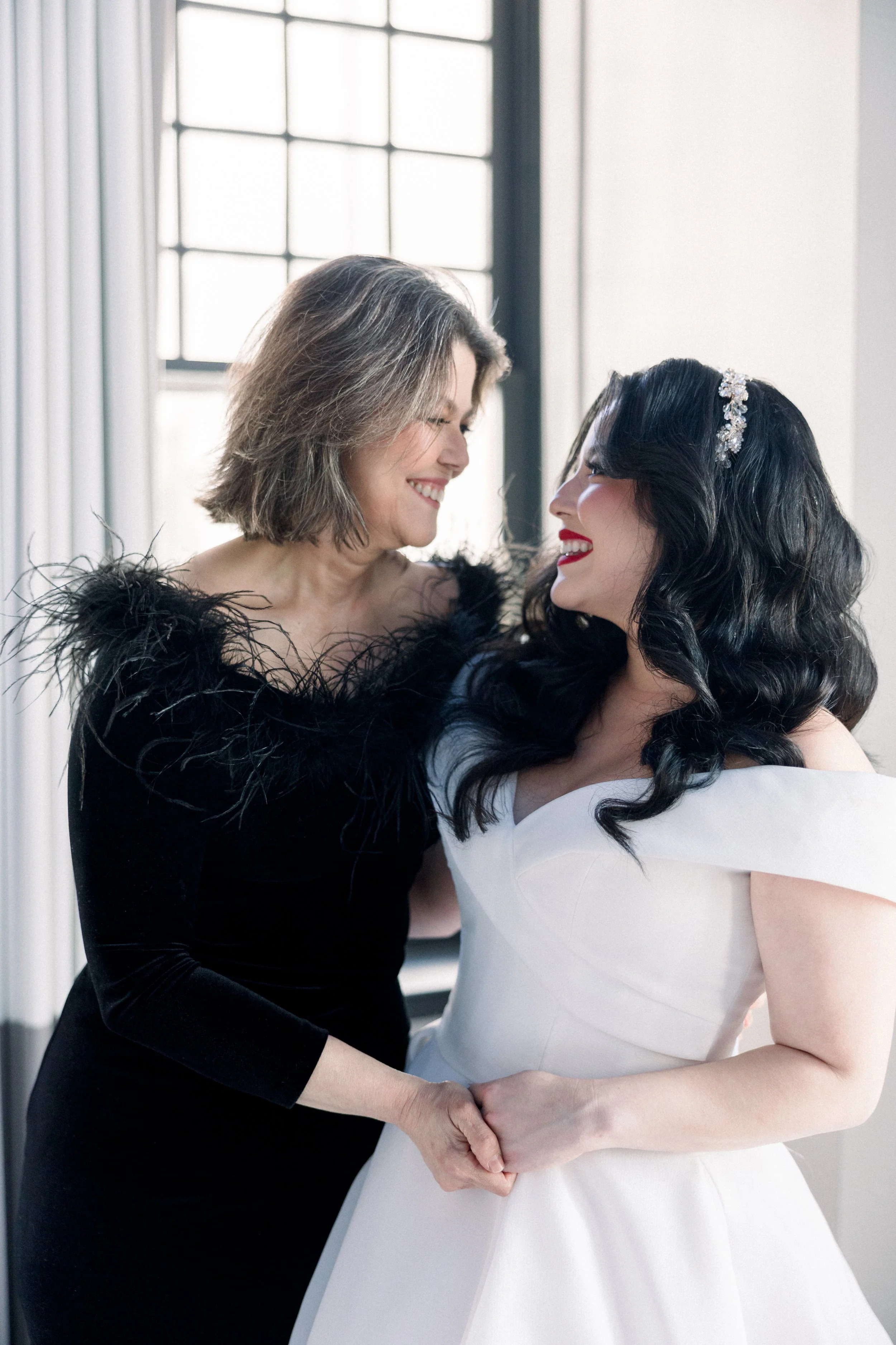 Two women, one in a black dress with feathered shoulders and the other in a white wedding gown, sharing an intimate moment and holding hands in front of a large window.