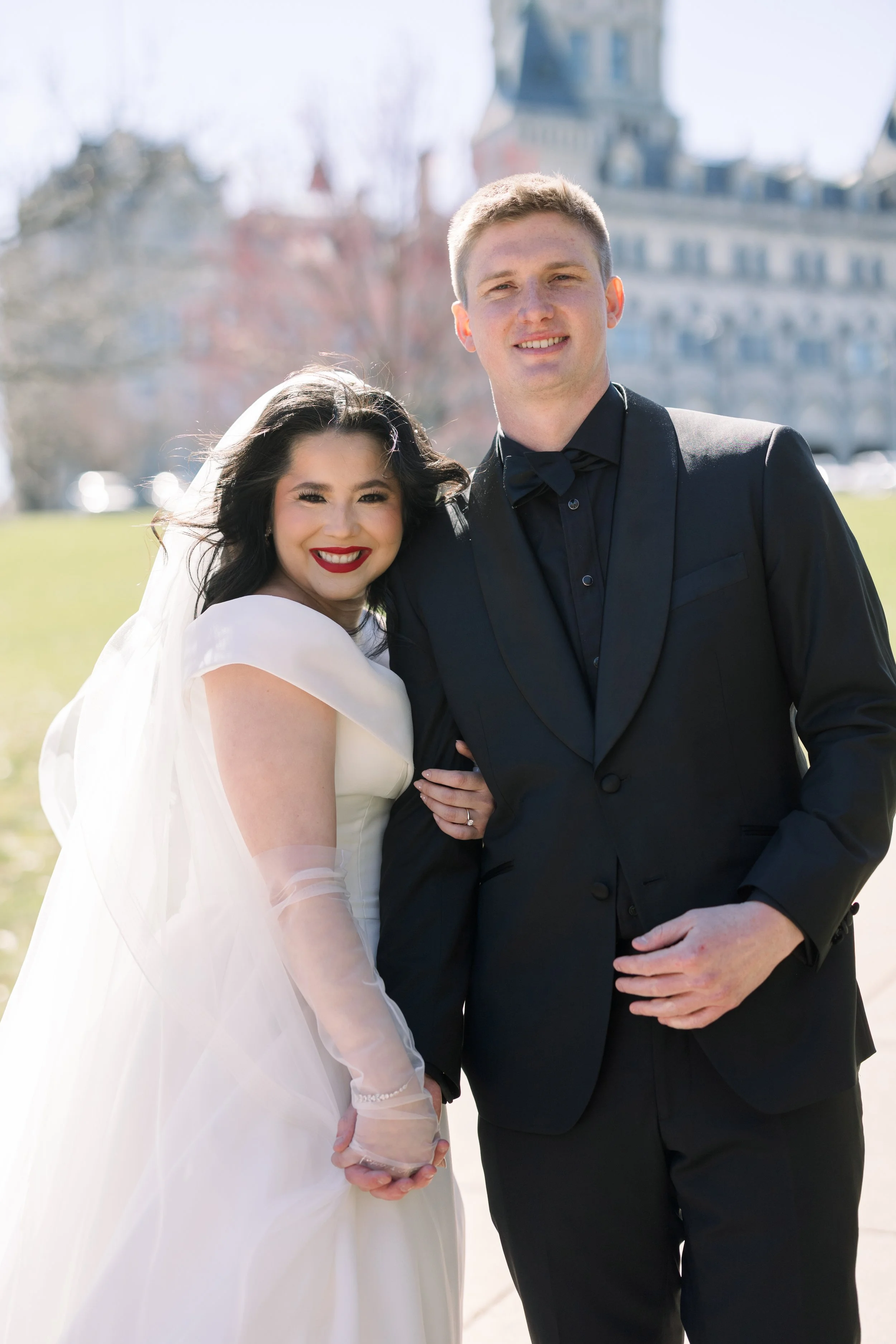 A smiling bride in a white wedding dress holding hands with a smiling groom in a black tuxedo, standing outdoors on a sunny day with a large building in the background.
