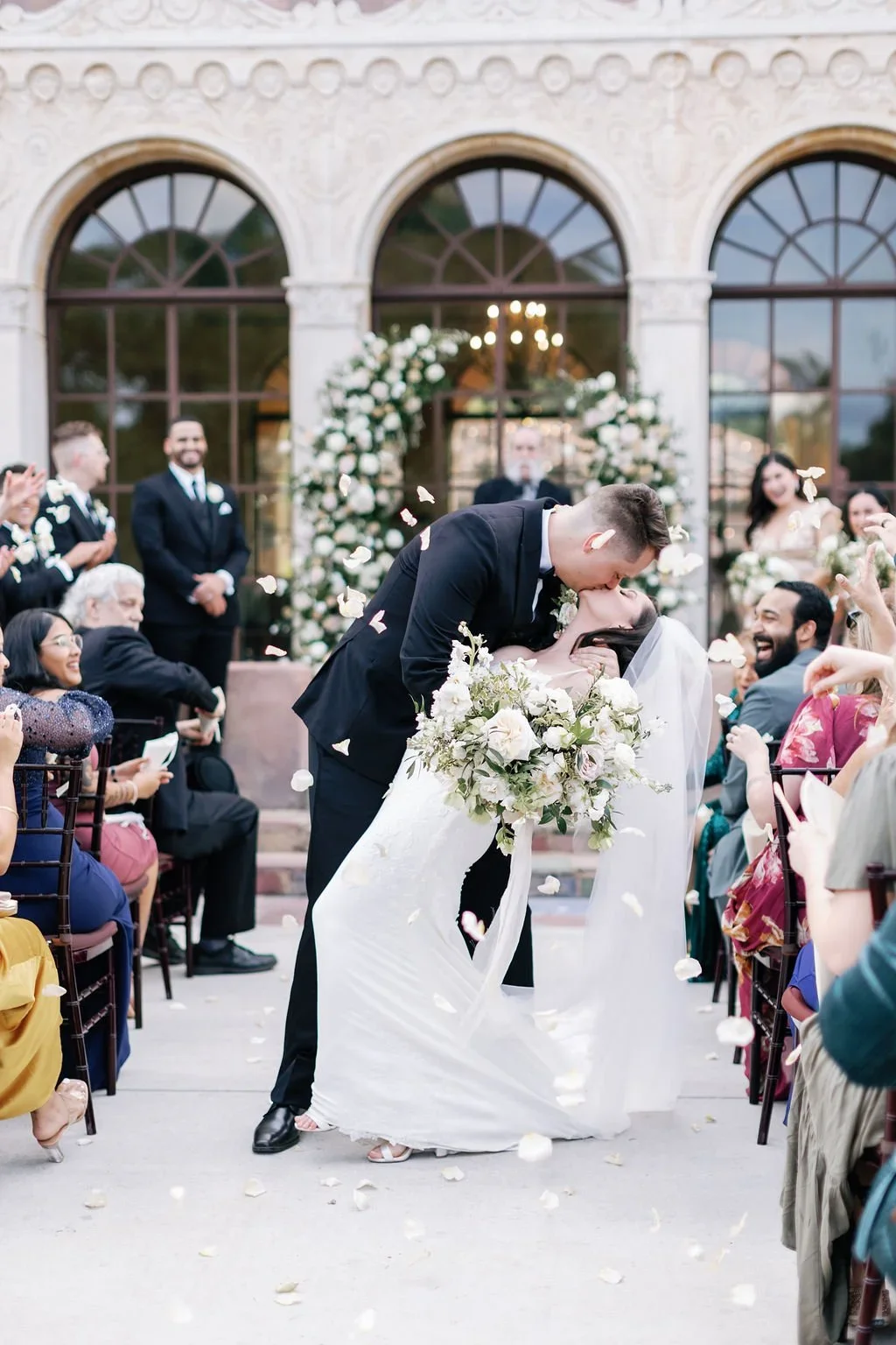 Couple sharing a kiss during their wedding ceremony outside a historic building with large arched windows, surrounded by friends and family throwing flower petals, with a floral arch in the background.