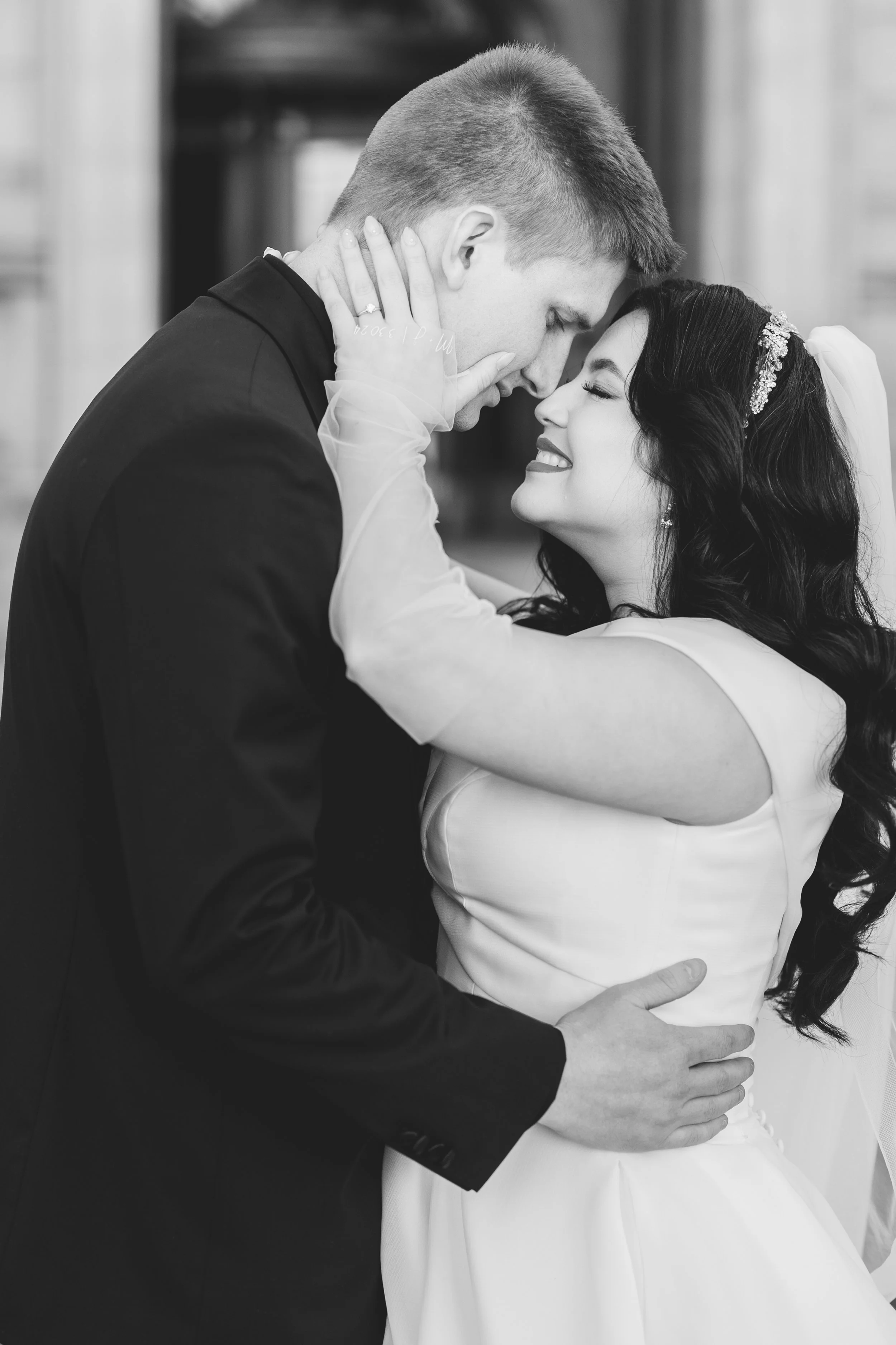 Black and white photo of a bride and groom during their wedding, with their foreheads touching and smiling, embracing each other.