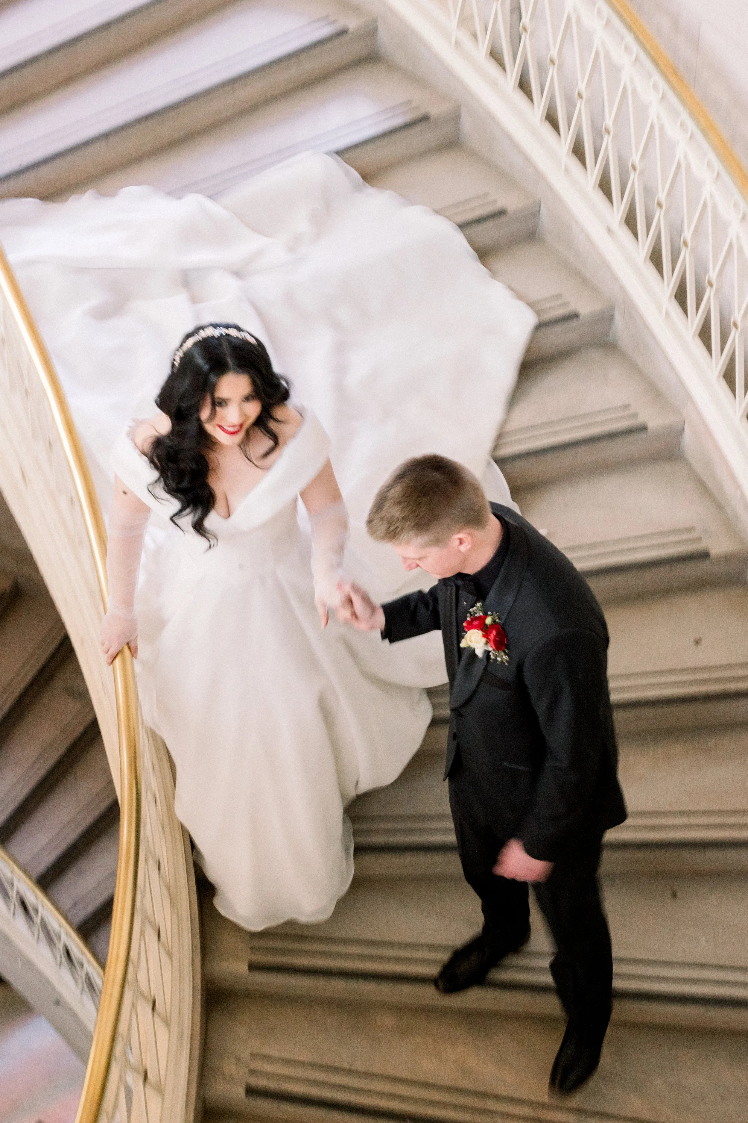 A bride and groom holding hands on a staircase, wedding attire, bride in a white gown and dark hair, groom in a black suit with a red boutonniere.