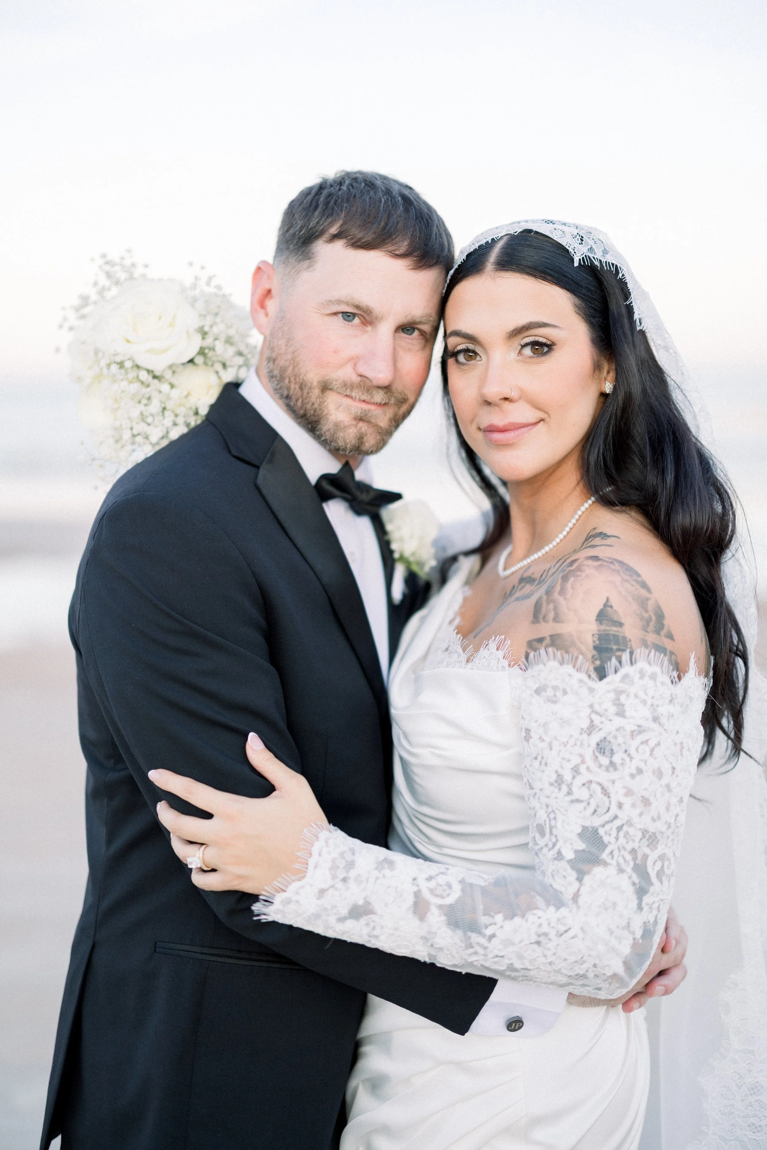 A bride and groom on their wedding day, standing close together outdoors with a blurred background, the bride with black hair and tattoos, wearing a lace wedding dress, the groom with a beard, wearing a black tuxedo, holding a bouquet of white flower