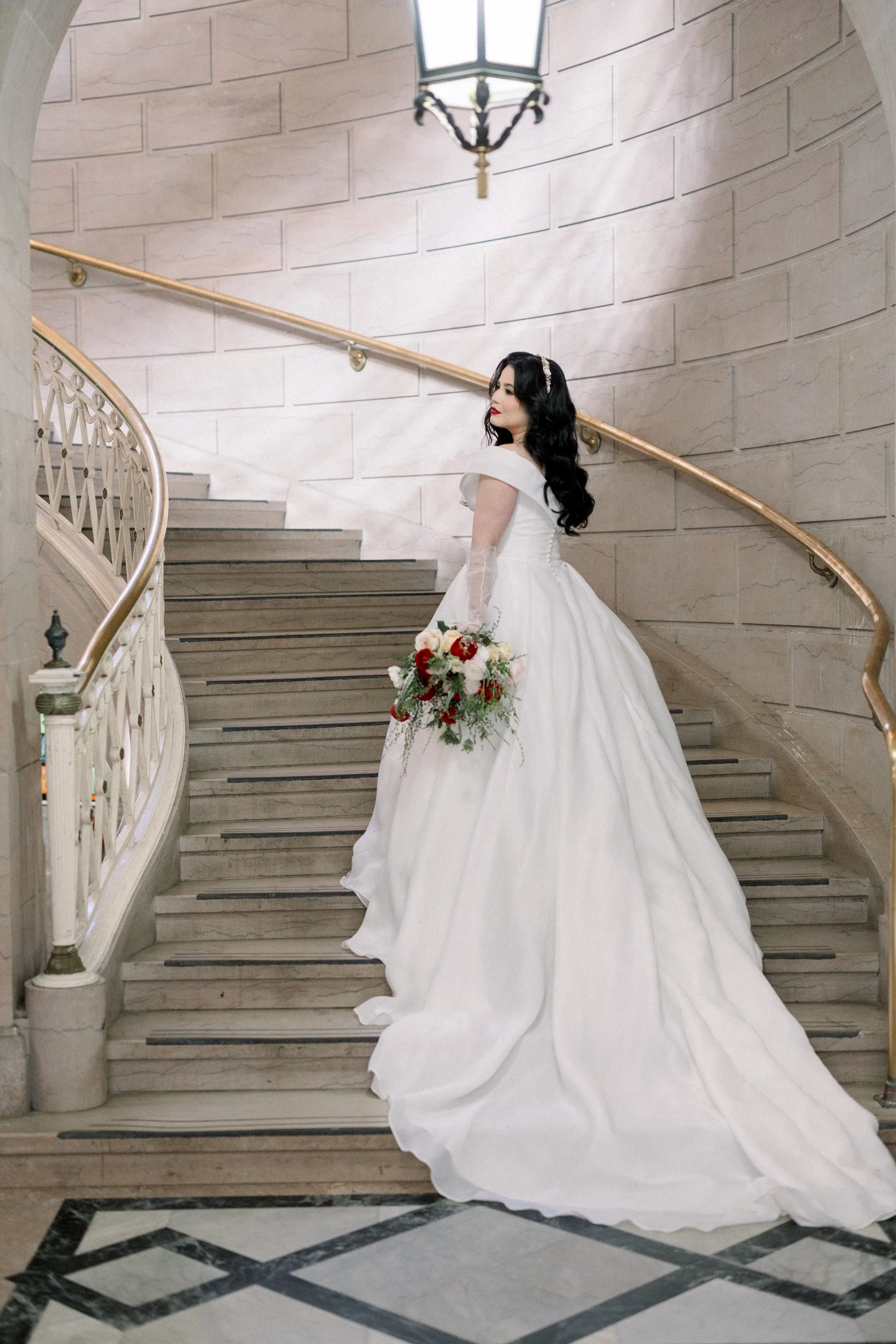 A bride in a white wedding gown holding a bouquet of flowers, standing on a staircase in an elegant building with stone walls and a decorative chandelier.