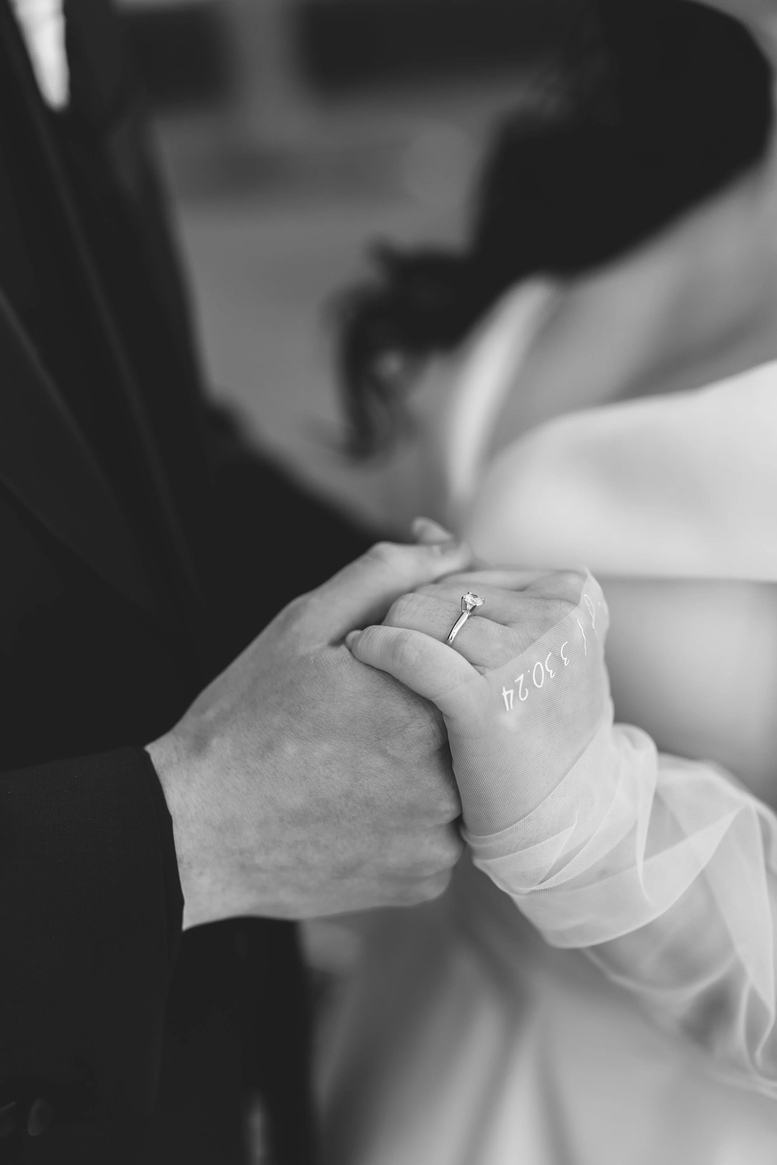 Black and white photo of a couple holding hands, showing a wedding ring on the woman's finger, with a blurred background of a woman in a wedding dress.