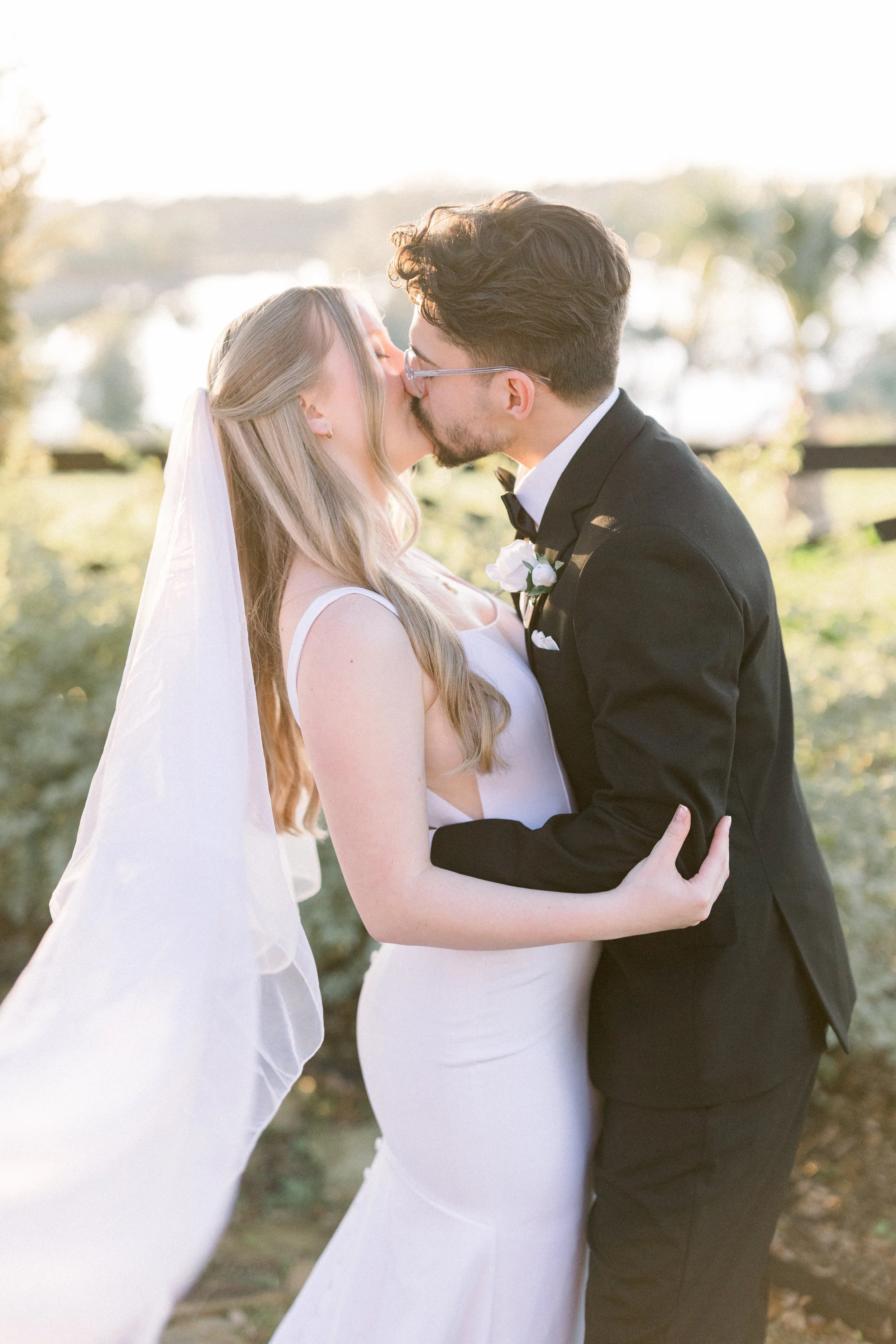 A newlywed couple sharing a kiss outdoors during sunset, the bride in a white wedding dress with a veil and the groom in a black tuxedo with glasses.