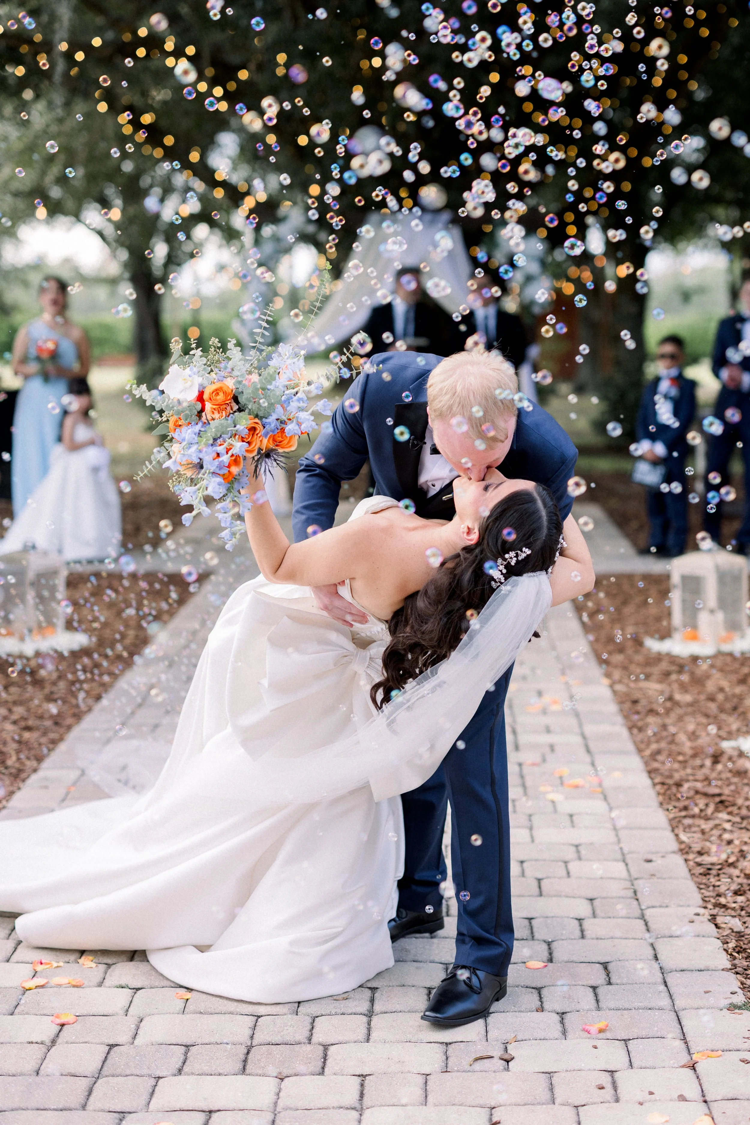 A newlywed couple shares a kiss under the shower of soap bubbles during their wedding ceremony outdoors. The bride is in a white dress holding a colorful bouquet, and the groom is in a dark suit. Bridesmaids and groomsmen are visible in the background, with trees and a pathway.