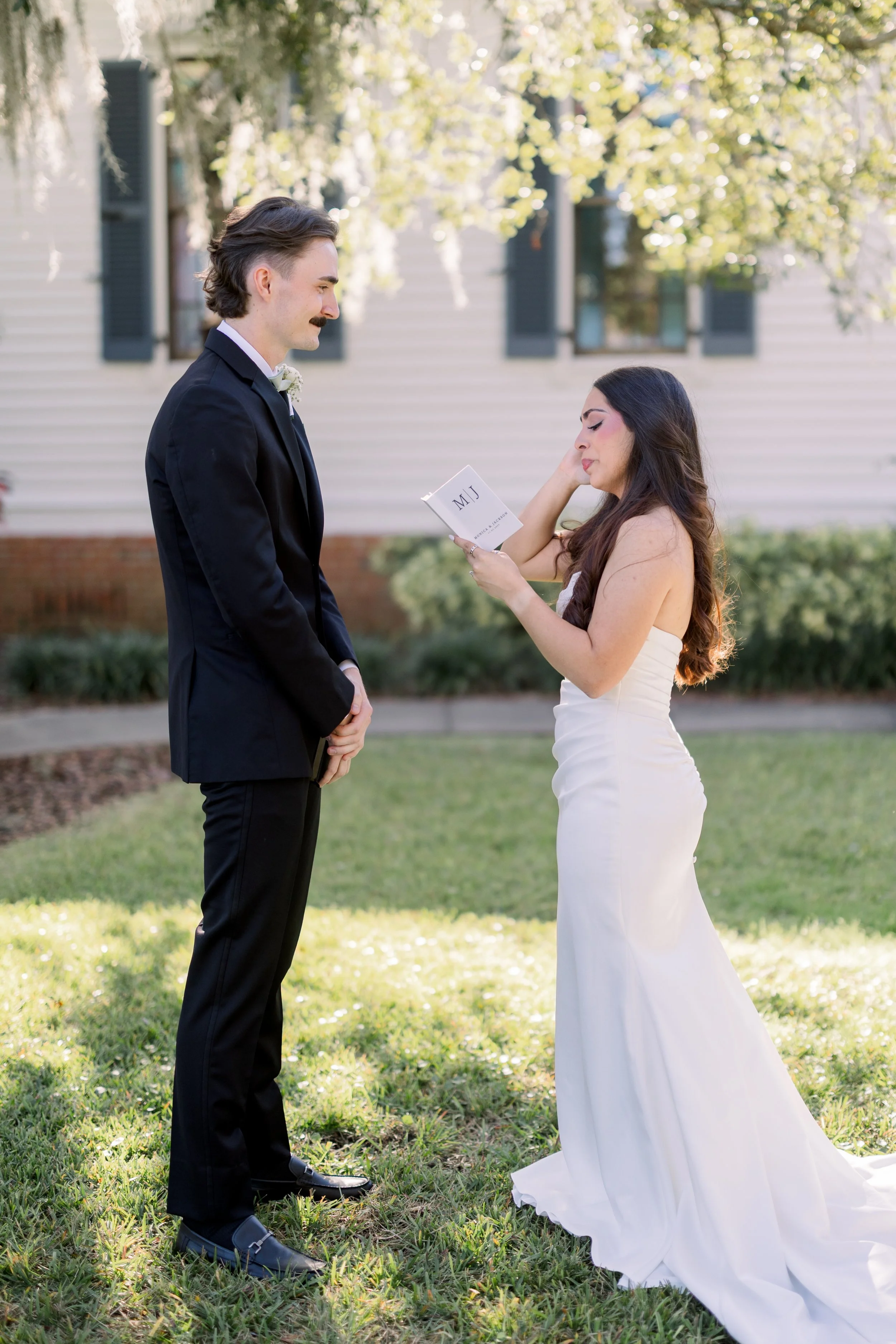 A bride and groom exchanging vows outdoors on a grassy lawn during daytime, with the bride reading vows from a small booklet, and trees and a house in the background.