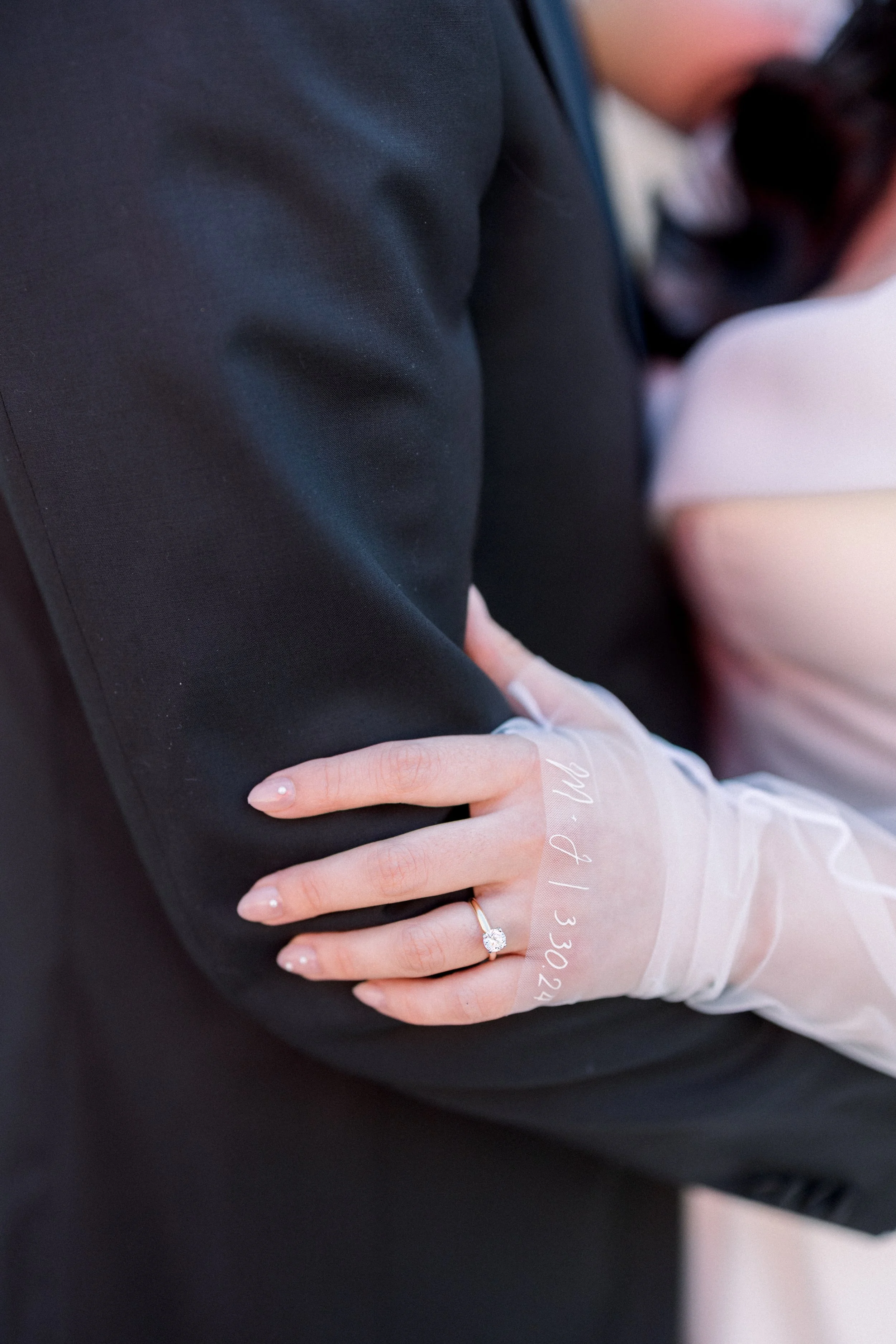 A woman wearing a wedding ring on her finger rests her hand on a man's arm at a wedding or formal event.