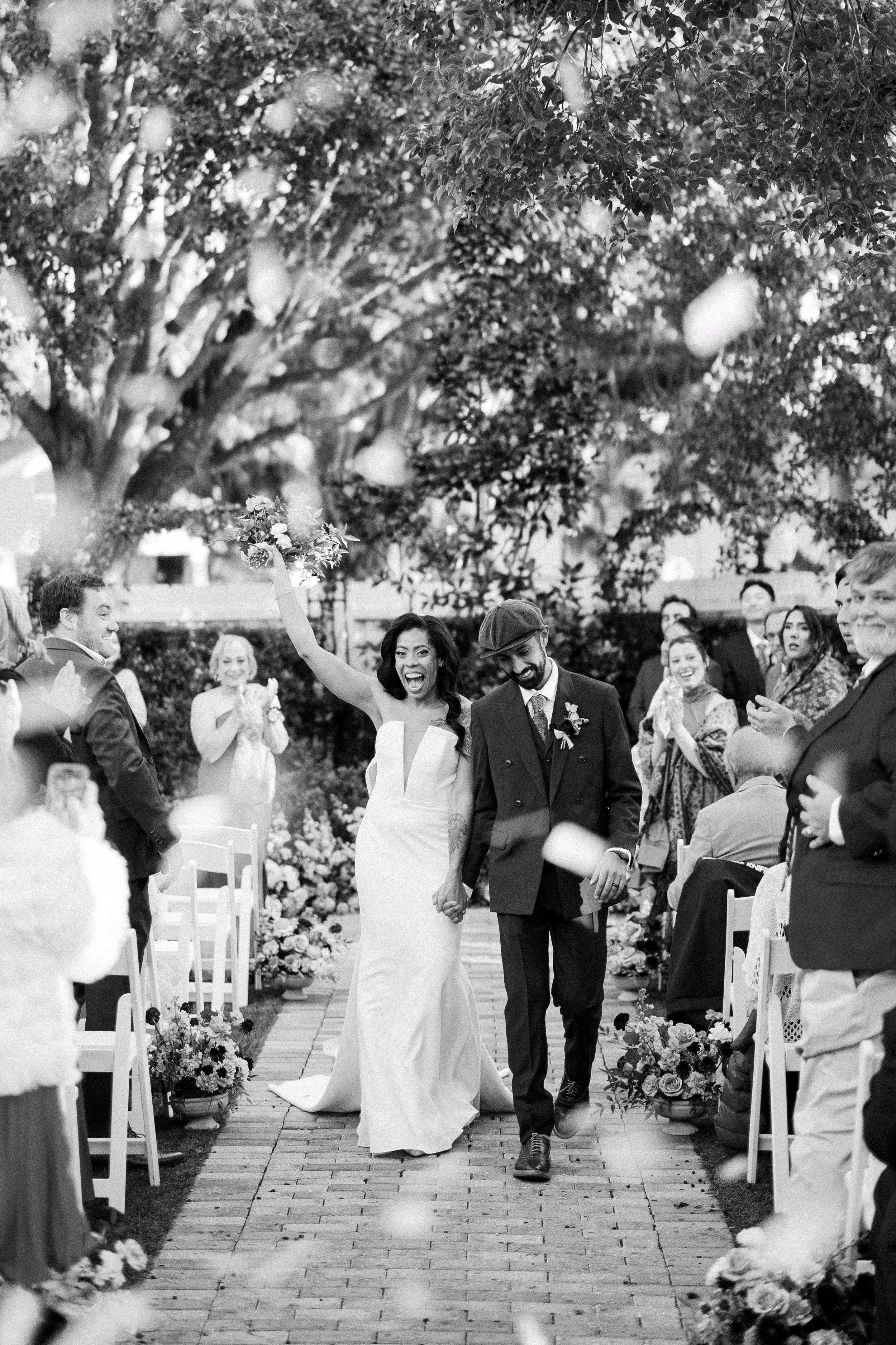 A bride and groom walking hand-in-hand down an outdoor wedding aisle with guests clapping and celebrating around them, surrounded by trees and floral decorations.