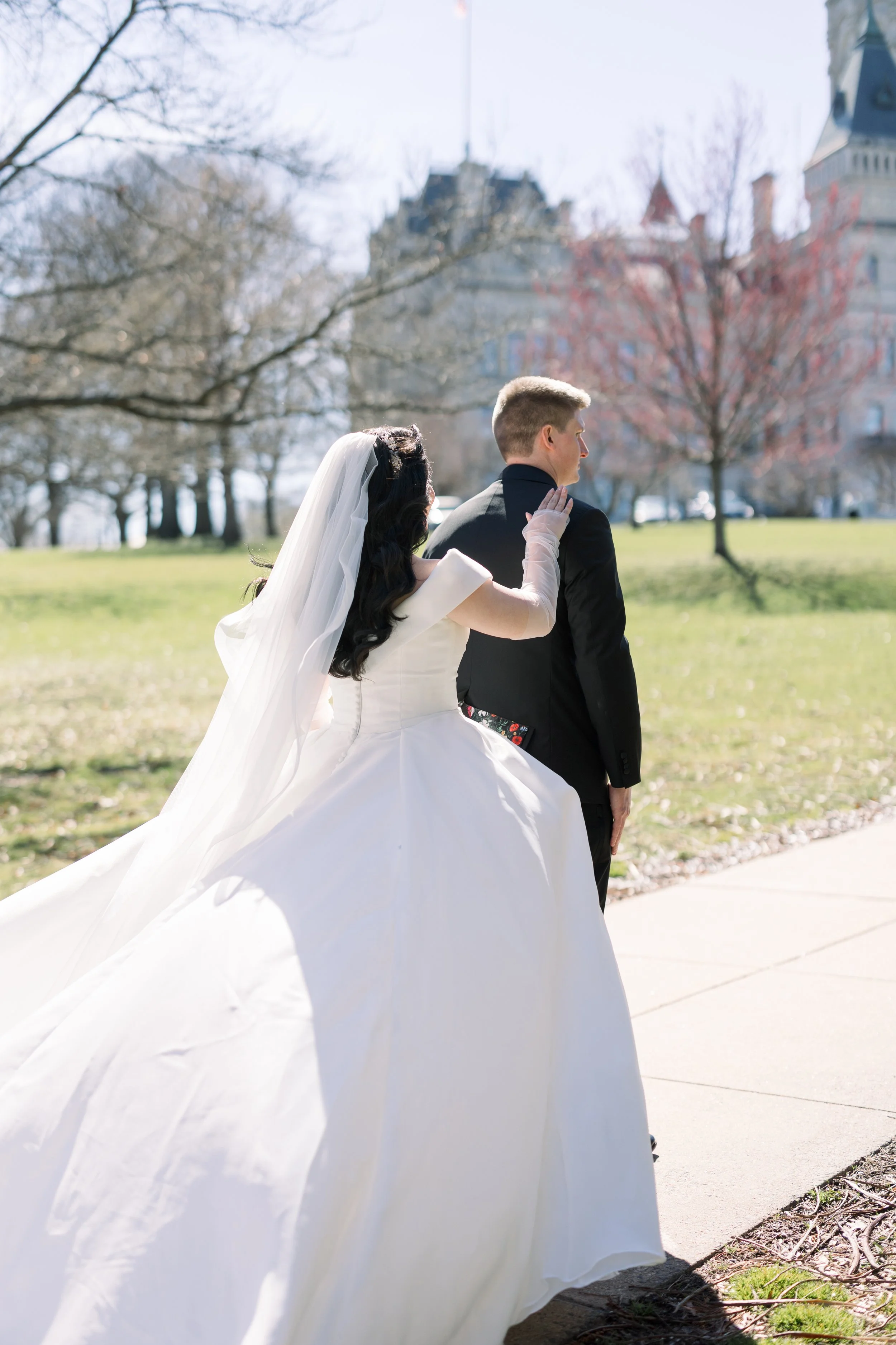 A bride and groom standing outdoors on a sunny day, with the bride adjusting the groom's jacket from behind, in a park setting with trees and a historic building in the background.
