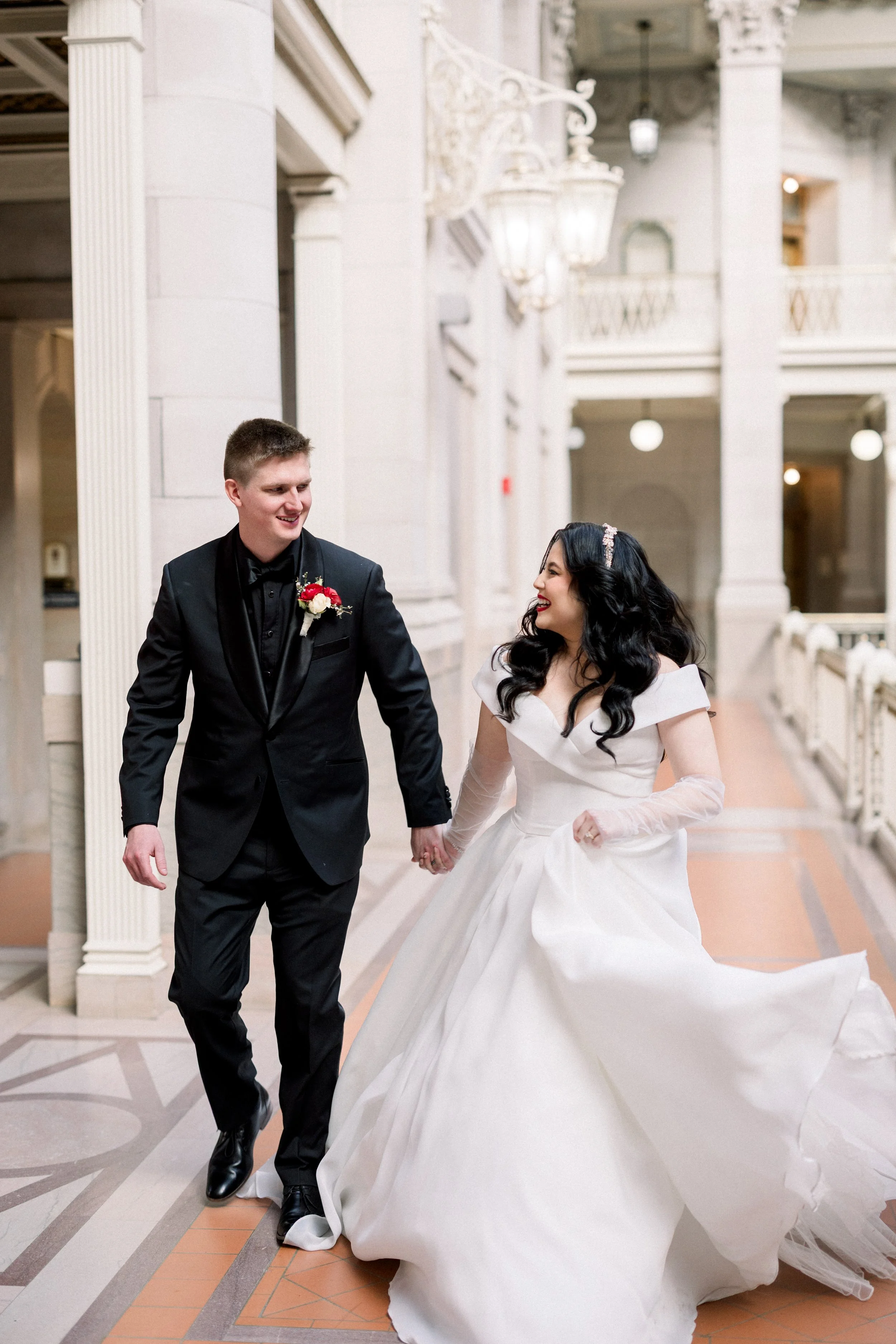 Bride and groom holding hands and smiling inside a grand, elegant building with white architectural details.