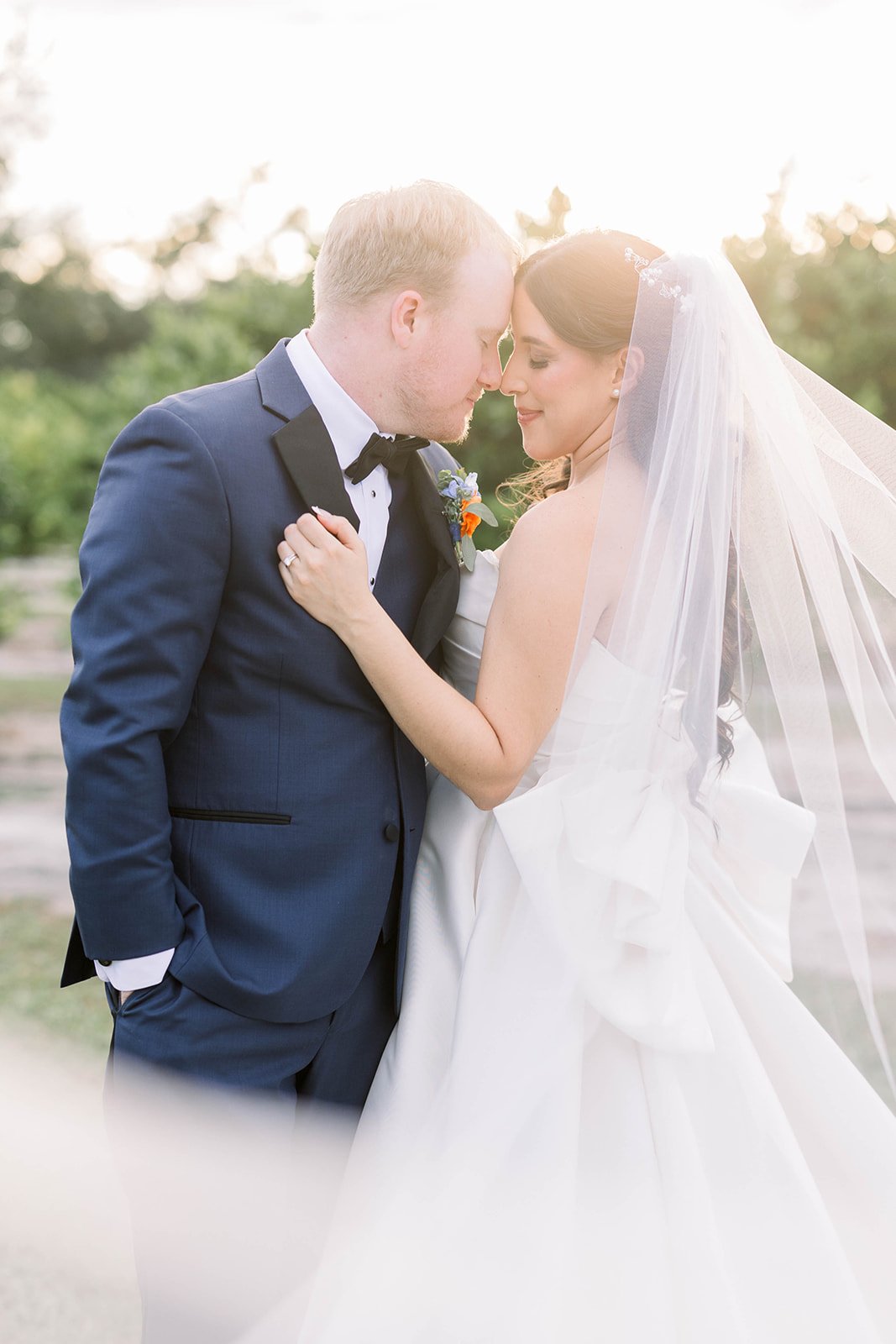 A newlywed couple embracing outdoors at sunset, with the groom in a navy tuxedo and the bride in a white wedding gown with a veil.