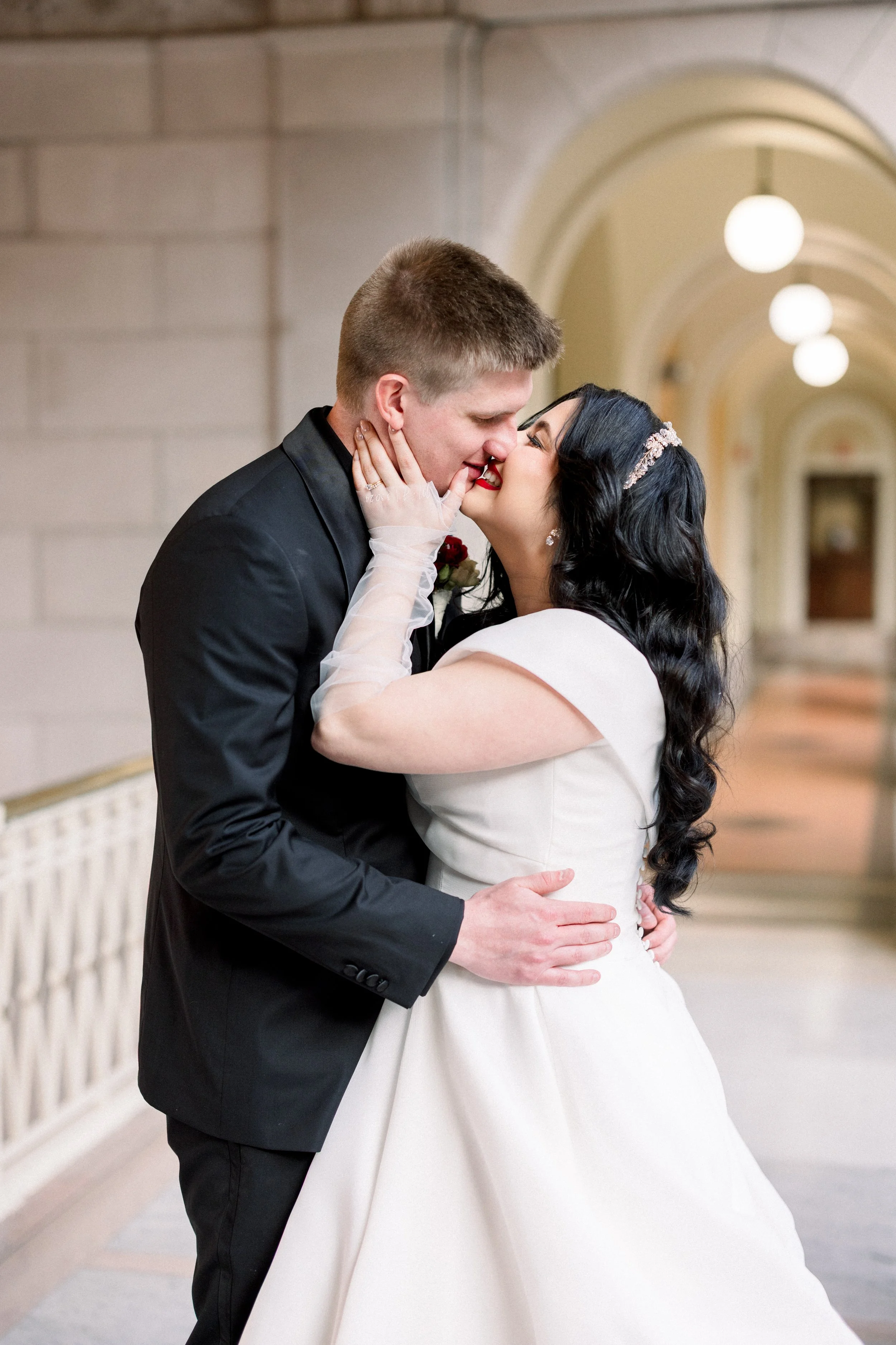 A newlywed couple sharing a kiss in a hallway. The groom is wearing a black suit, and the bride is in a white wedding dress with dark curly hair, wearing earrings and a hair accessory.