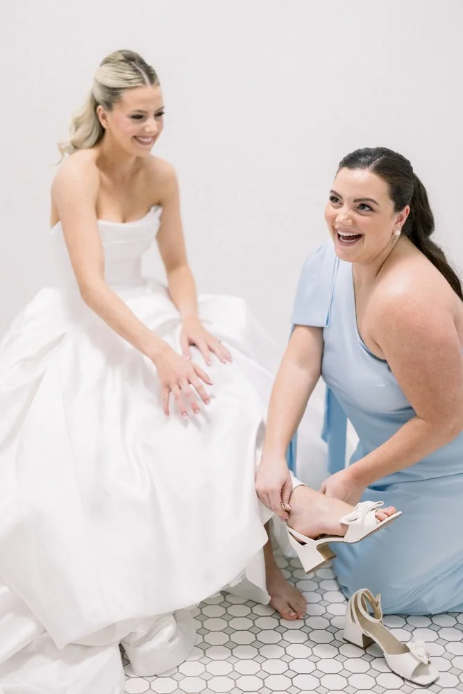 Bridesmaid helps the bride put on her wedding shoes as they laugh together while getting ready