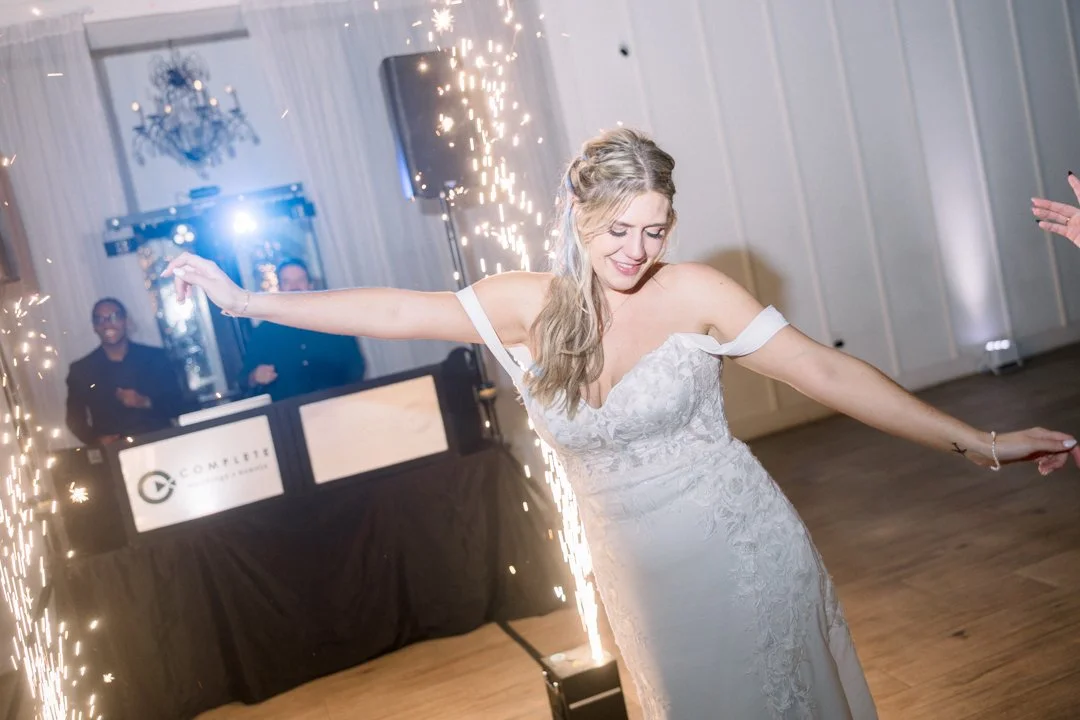 A bride in a white wedding dress dancing with sparklers at her wedding reception, with a DJ booth and guests in the background.