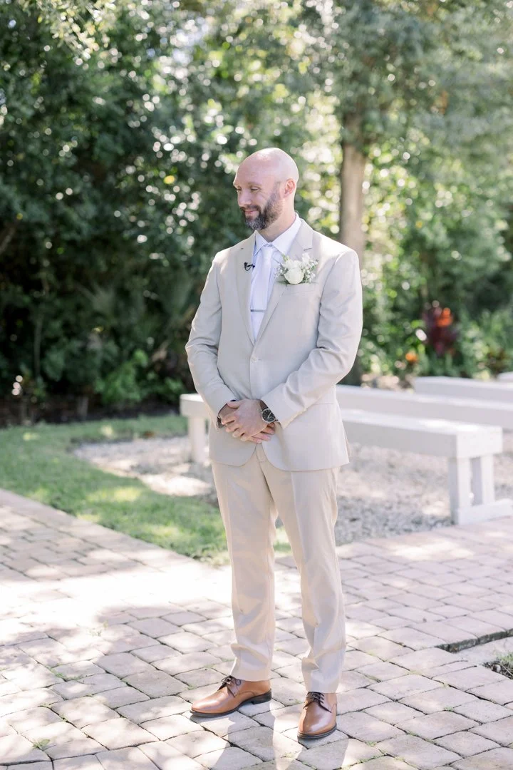 A man in a cream-colored suit standing outdoors on a brick pathway, with trees and greenery in the background. He has a beard, is bald, and is wearing a white shirt, a boutonniere, and brown shoes.