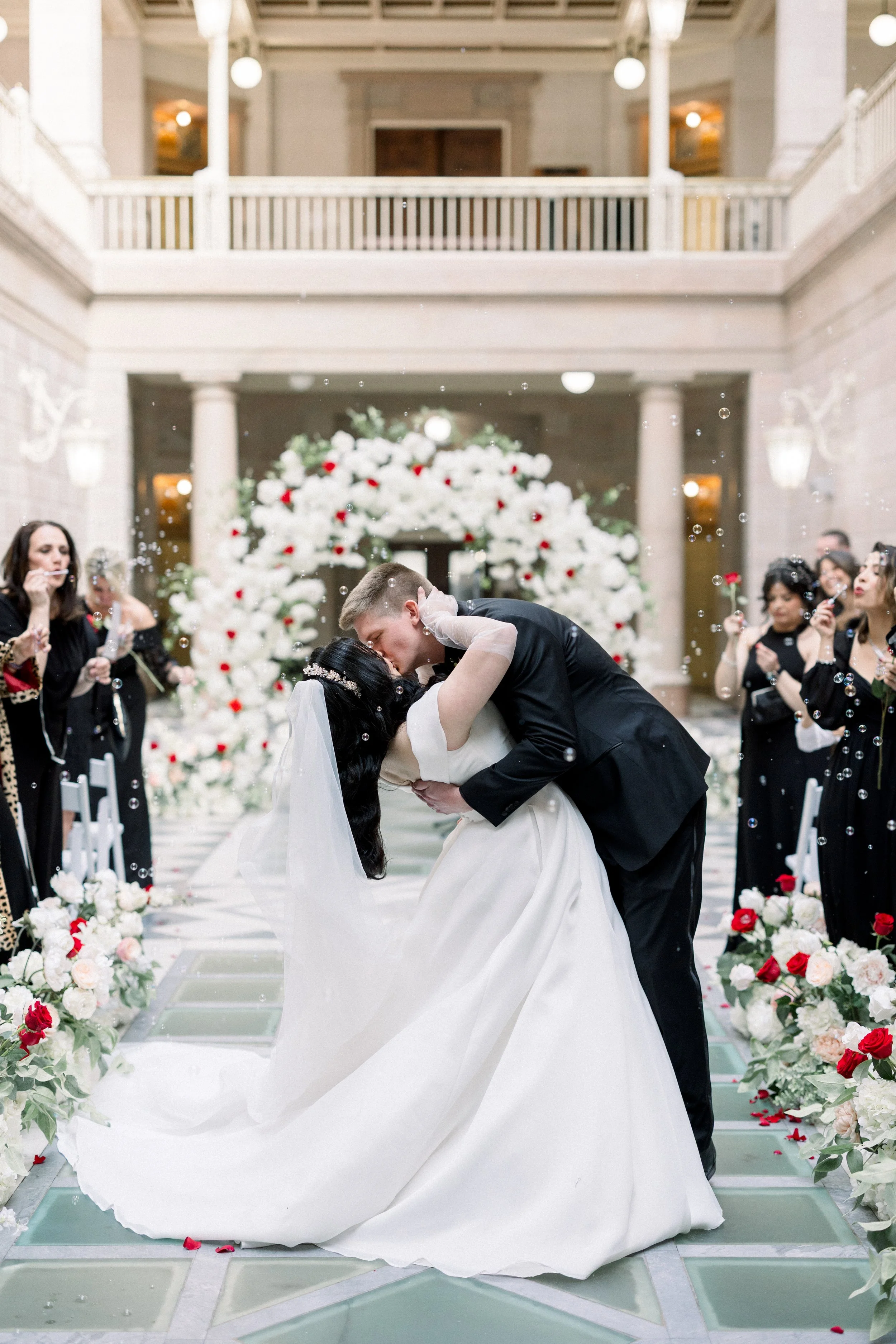 Bride and groom kissing during their wedding ceremony in an elegant hall decorated with white and red flowers, surrounded by friends and family.