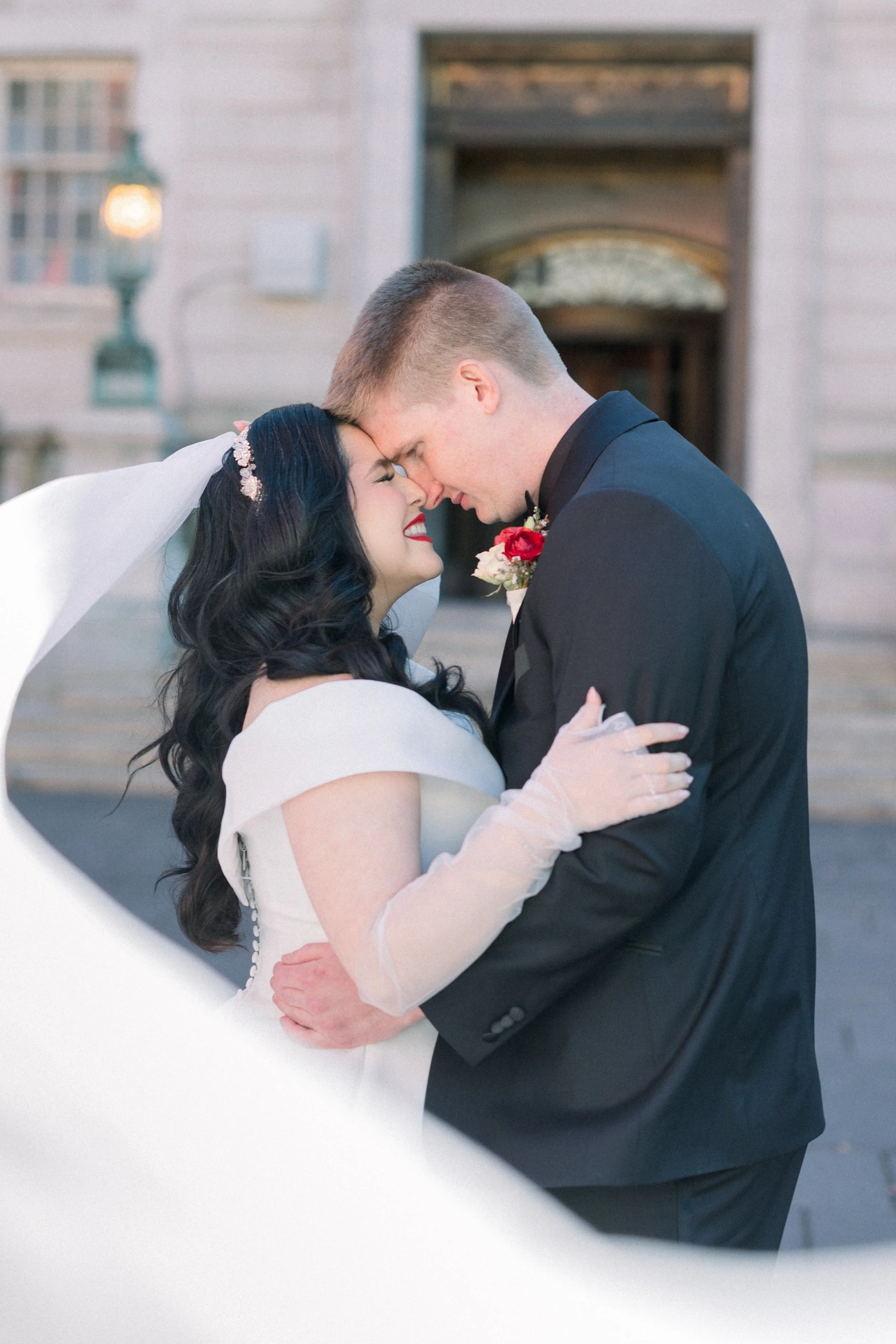 A bride and groom embrace closely with their foreheads touching during their wedding, outside in front of a building with an open door and vintage lamp, sharing a romantic moment.