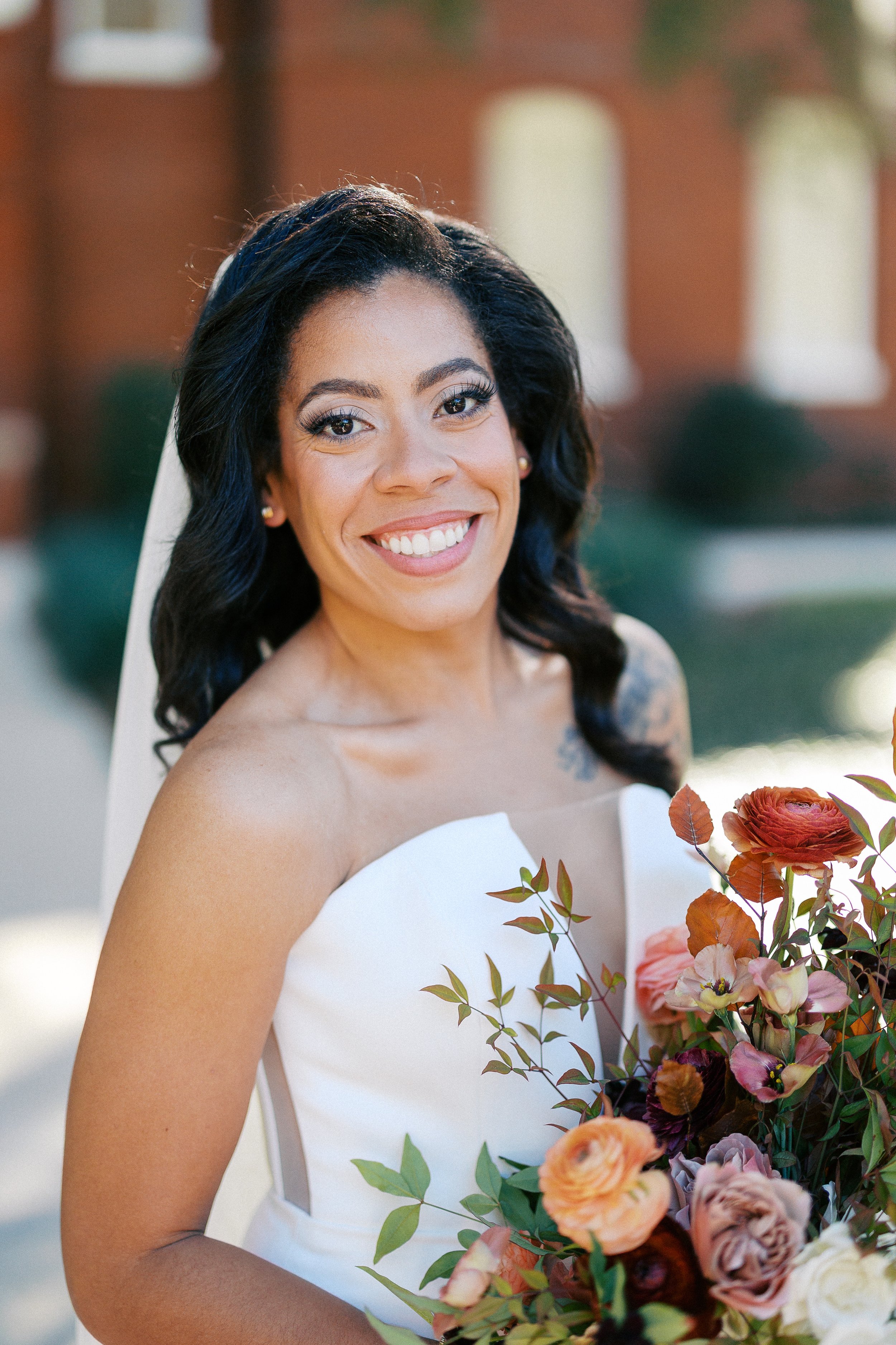 Smiling bride in a white strapless wedding dress holding a bouquet of pink, peach, and burgundy flowers with green leaves, outdoors in front of a brick building.