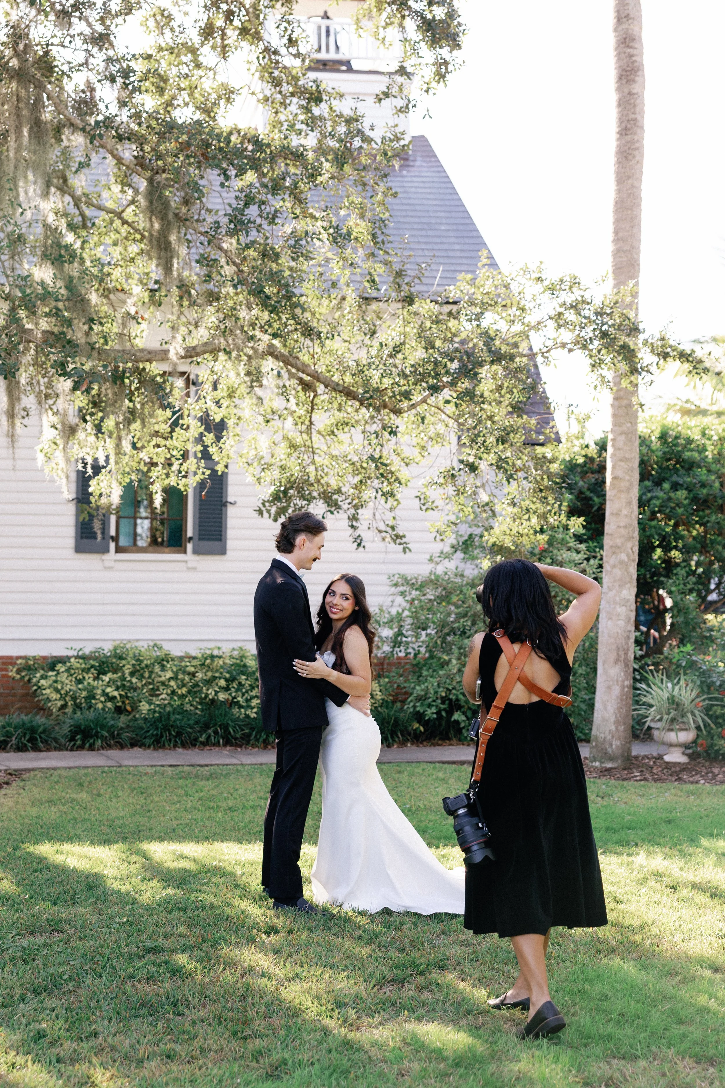 A couple dressed in wedding attire standing on a grassy lawn, being photographed by a woman with a camera. The couple is smiling and embracing, with a house and trees in the background.