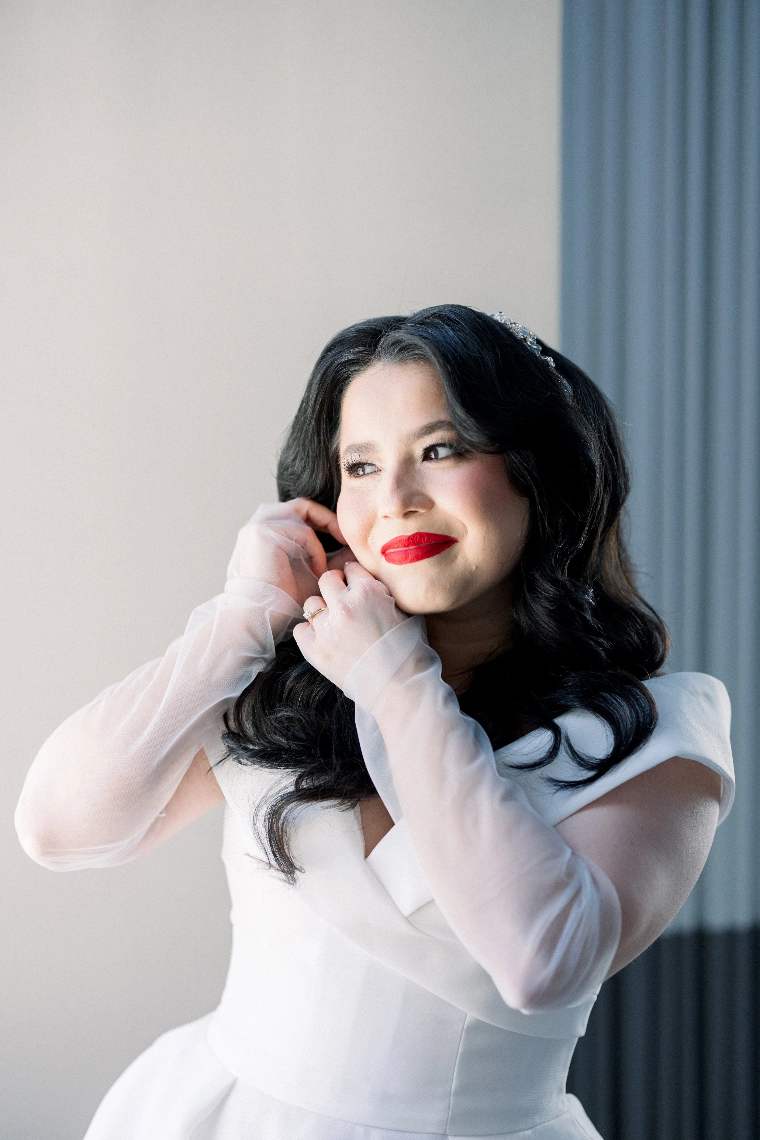 Young woman with black wavy hair and red lipstick, wearing a white dress and sheer gloves, smiling softly while adjusting her earrings.