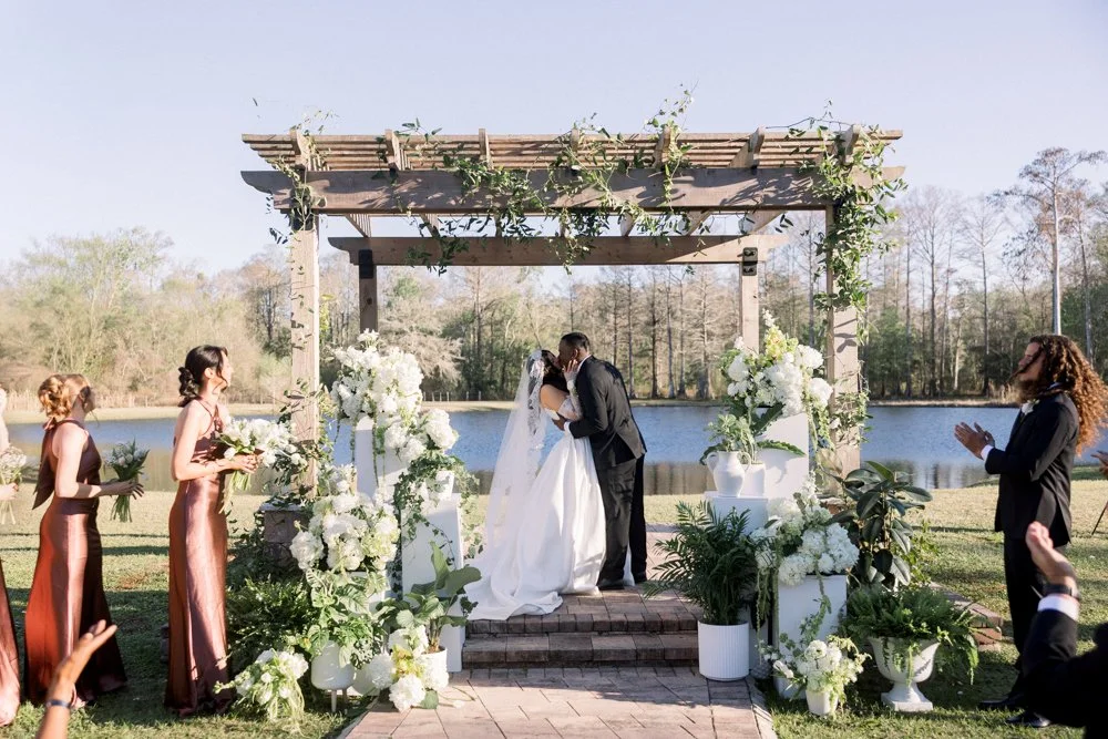 Bride and groom kiss under a flower-covered pergola during a lakeside Central Florida wedding ceremony, photographed by Created by Patty