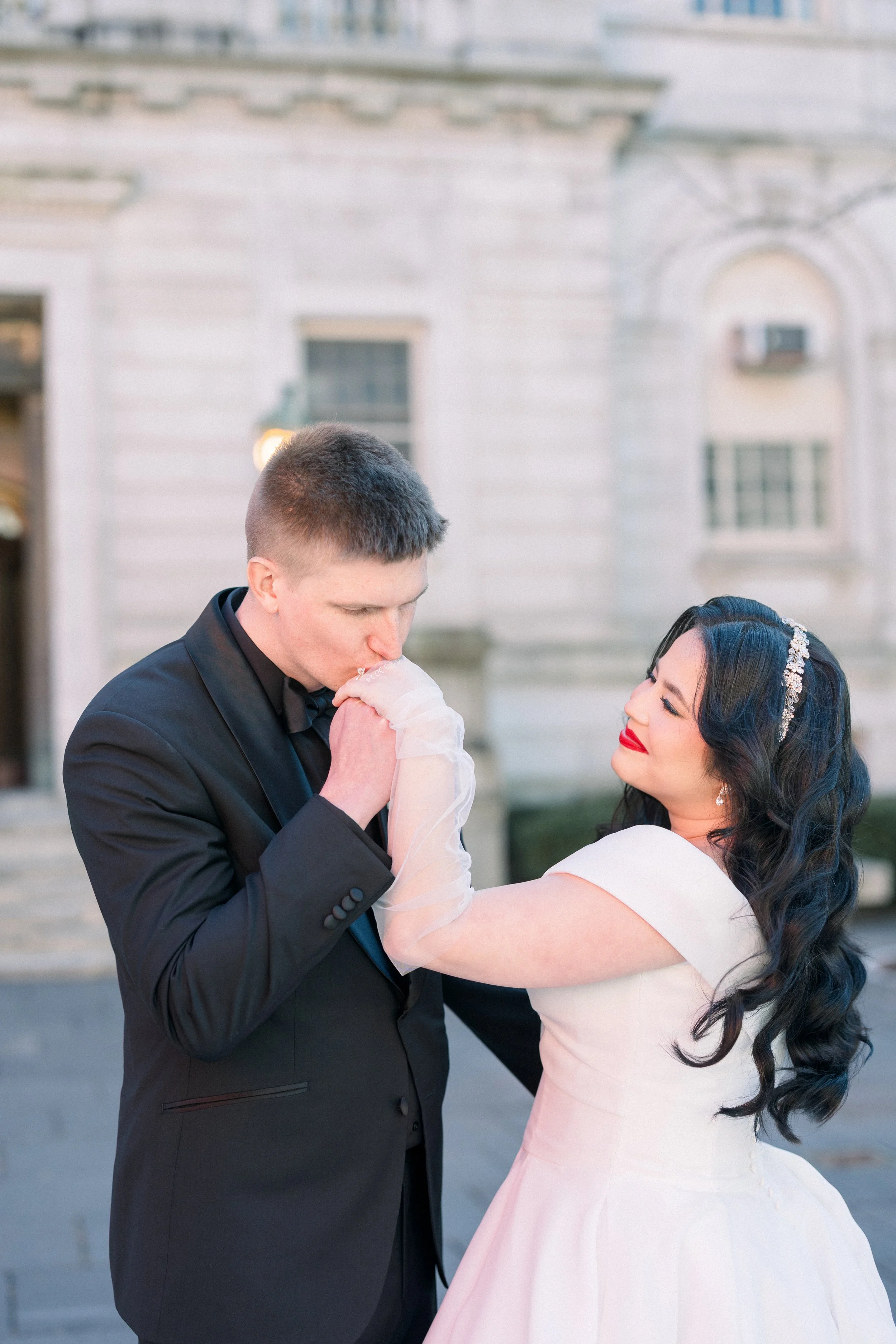 A man in a black tuxedo kisses a woman in a pink dress on her hand during a dance or romantic moment outside a historic building with stone walls.