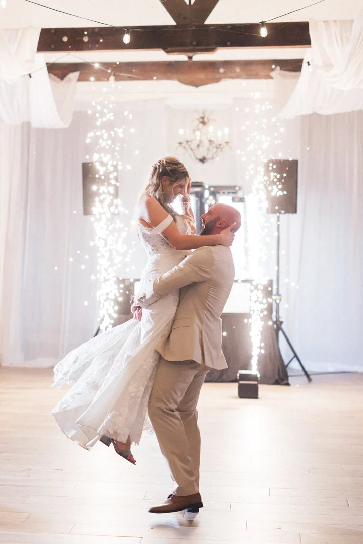 A couple in formal wedding attire dancing, with the man lifting the woman off the ground, surrounded by sparklers and string lights in a decorated indoor venue.