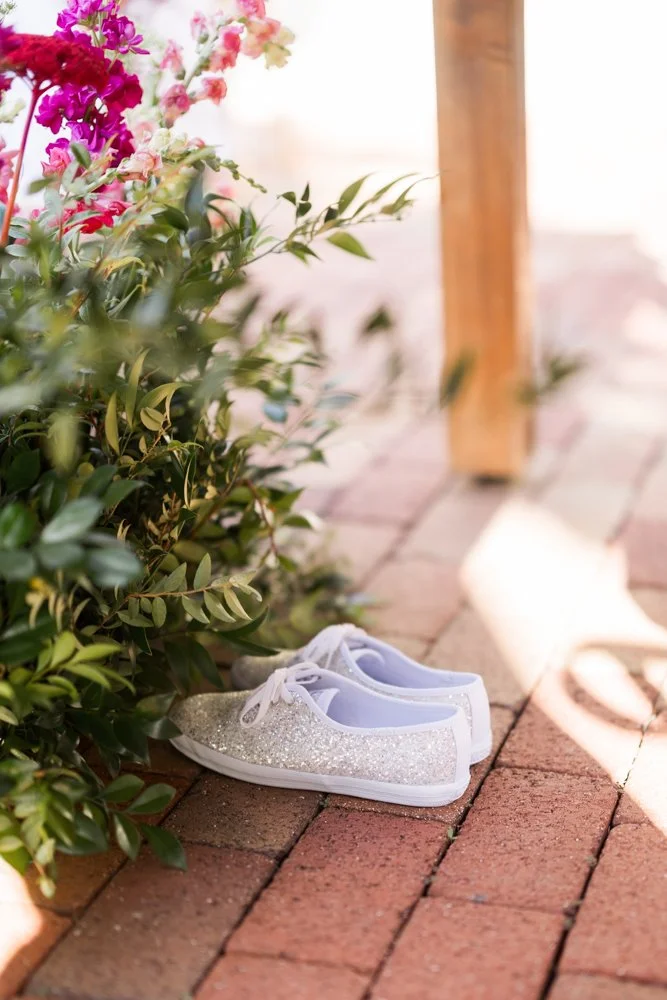 White wedding sneakers beside bright pink florals on a brick patio, wedding detail photo by Created by Patty