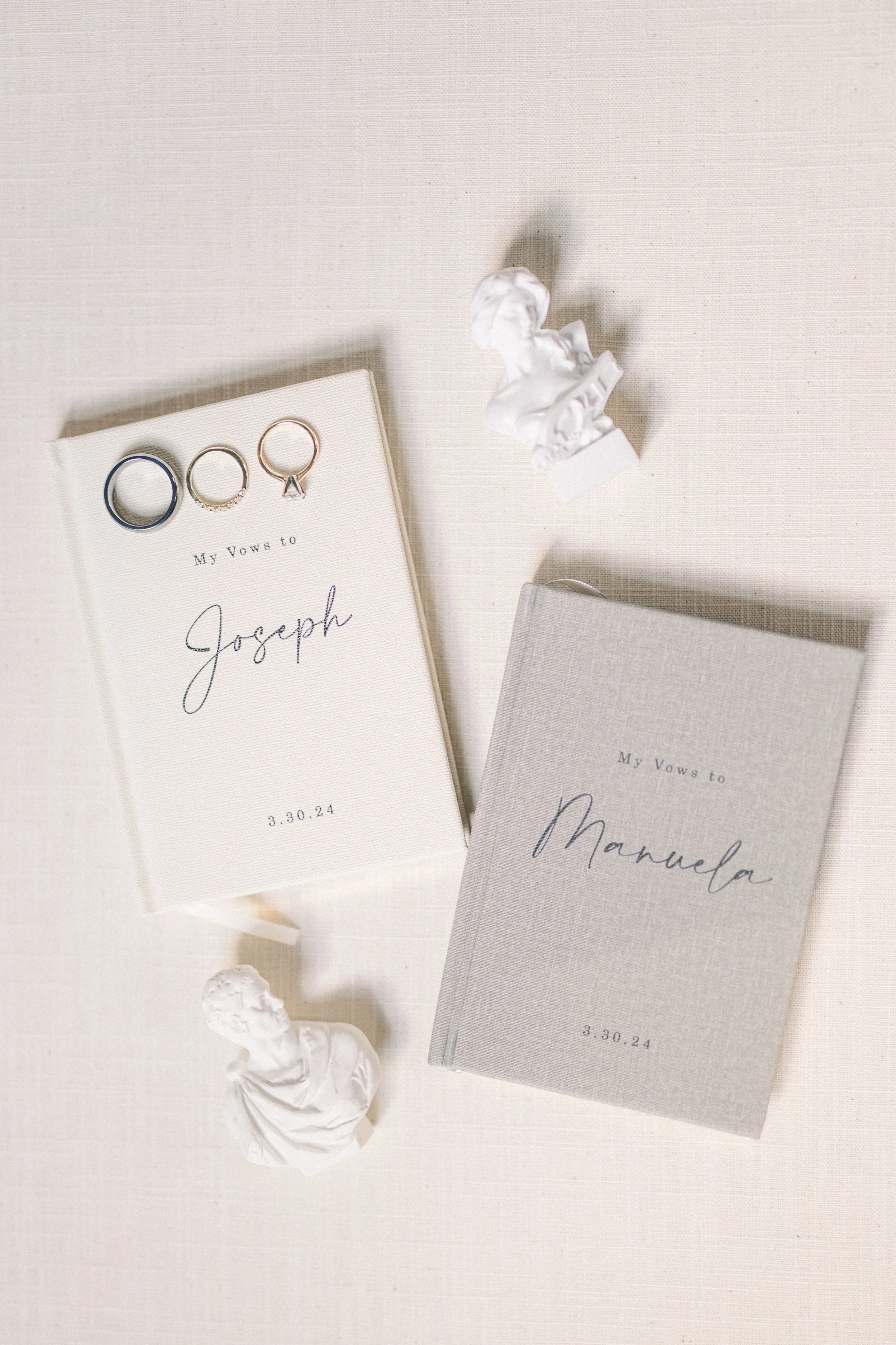A flat lay of two hardcover vow books, one titled 'My Vows to Joseph' with a date of 3.30.24, and the other titled 'My Vows to Manuela' with the same date. There are three rings placed on Joseph's vow book, and two small white bust sculptures, one of