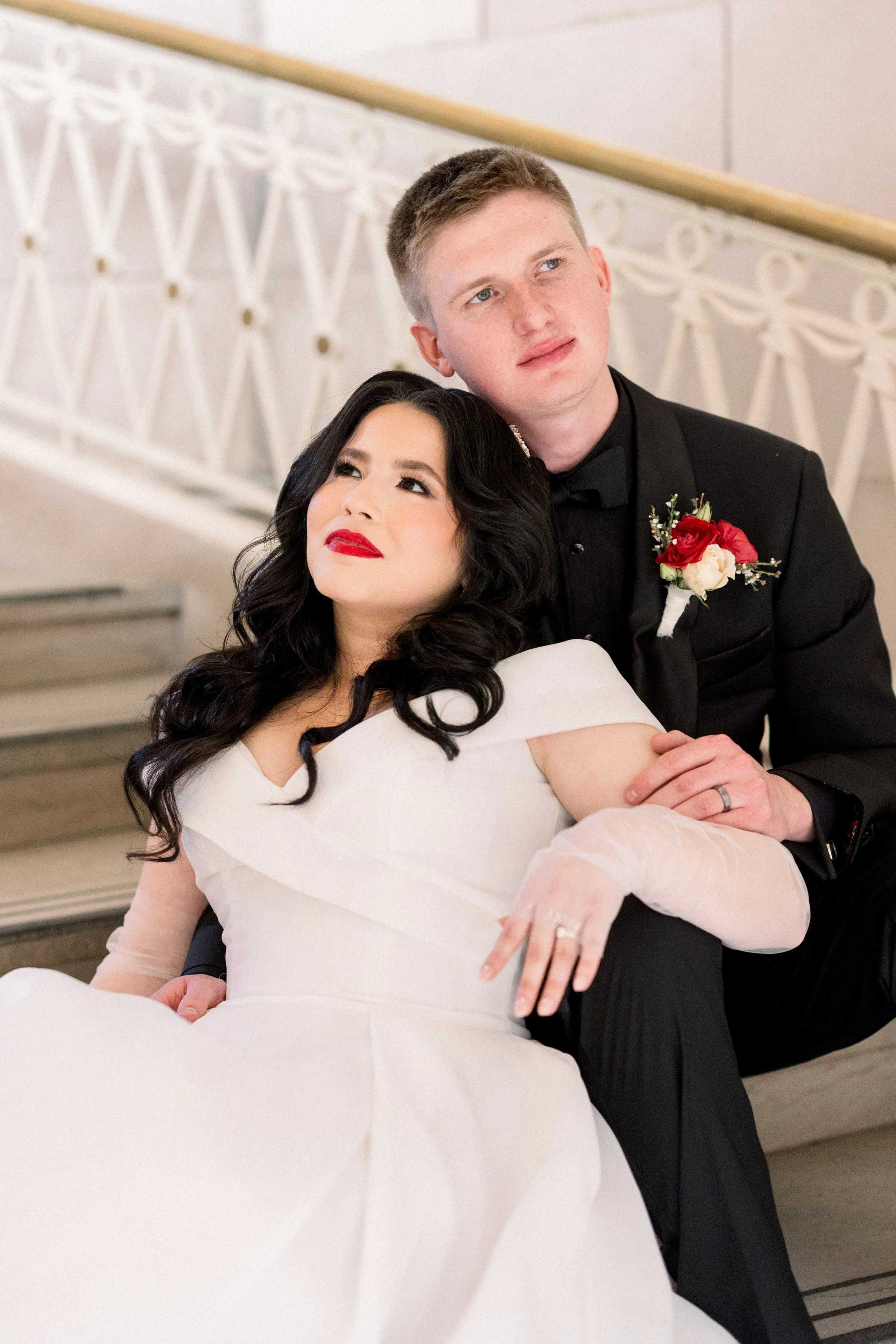 A newlywed couple on a staircase, with the bride sitting and the groom kneeling behind her, close together, dressed in wedding attire. The bride has dark hair and red lipstick, and the groom is wearing a black tuxedo with a red and white boutonniere.