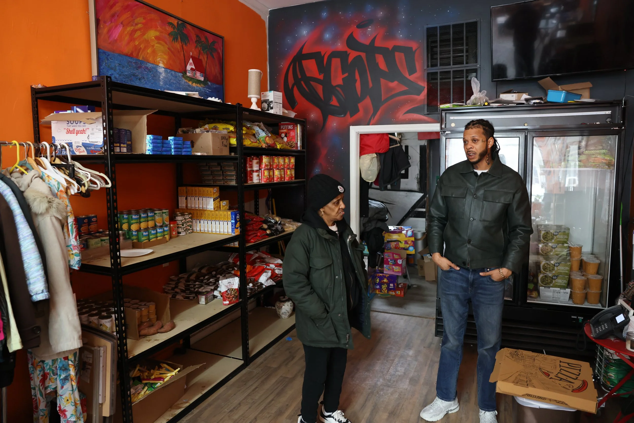 Two people, a woman and a man, are inside a store talking. The woman is wearing a green jacket and a black beanie, and the man is wearing a black leather jacket and jeans. Shelves behind them are stocked with canned foods, boxed goods, and snacks. The store has colorful wall art, including a sunset scene and graffiti-style designs.