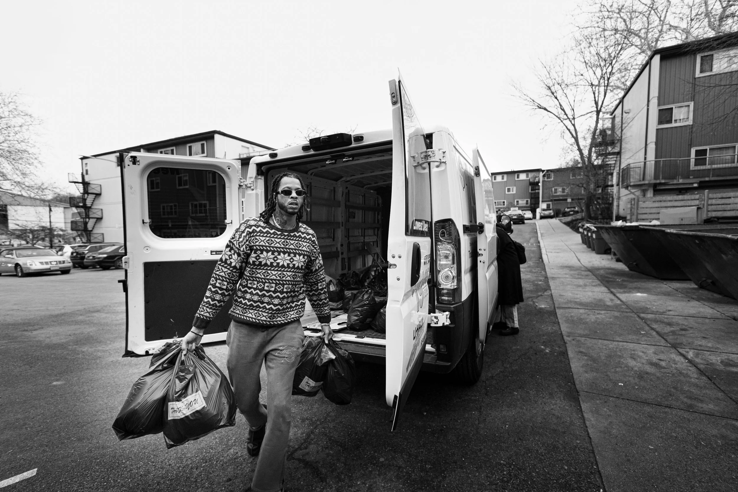 A man with dreadlocks wearing sunglasses, a patterned sweater, and loose pants, carrying black grocery bags, standing outside near an open delivery van with residential buildings and parked cars in the background.