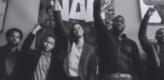 Group of five men standing with fists raised, smiling, in a room with artwork and a window.