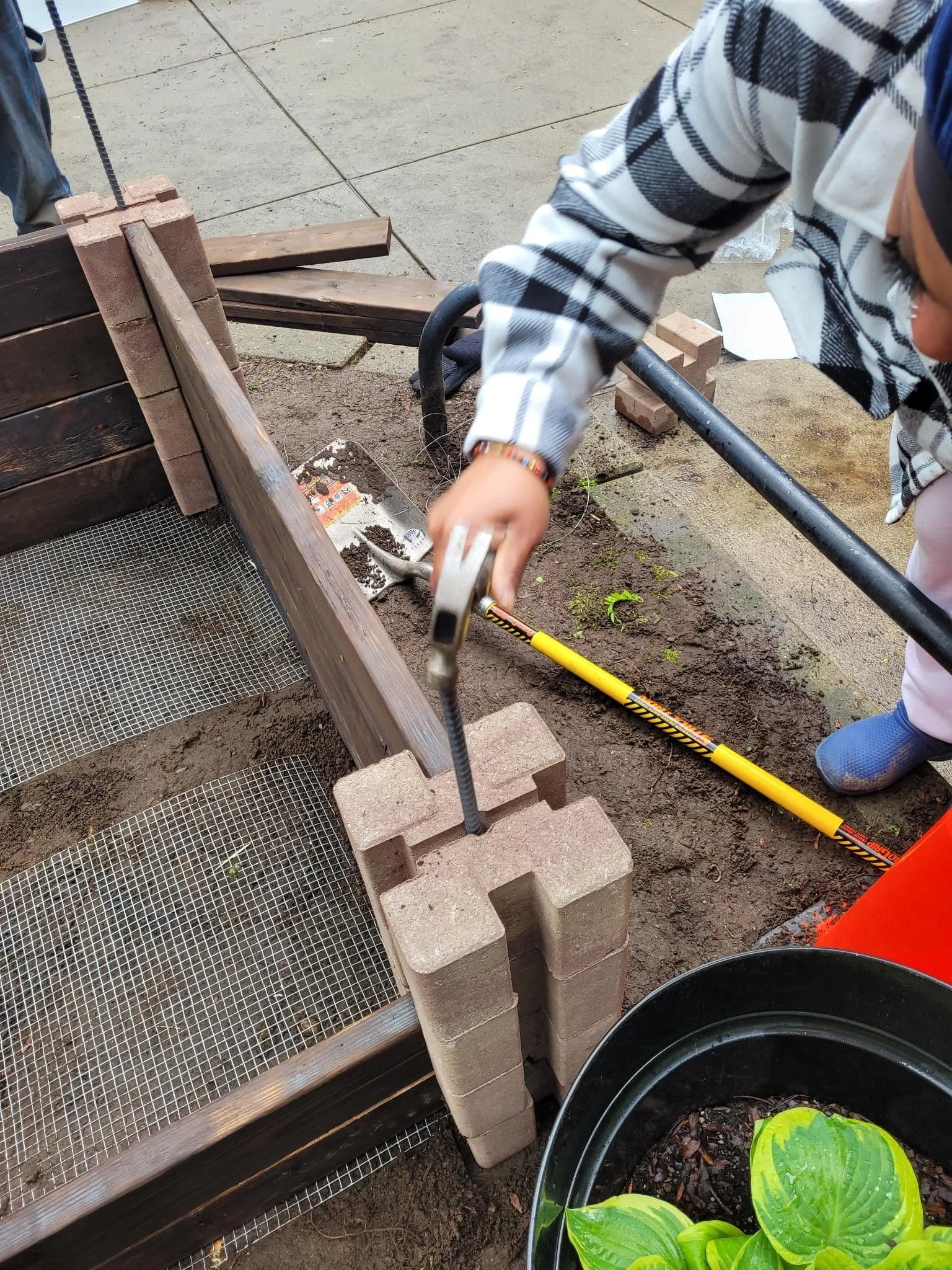 A person working on a garden project, laying bricks or blocks alongside a wooden frame or garden bed, with gardening tools and plants nearby.