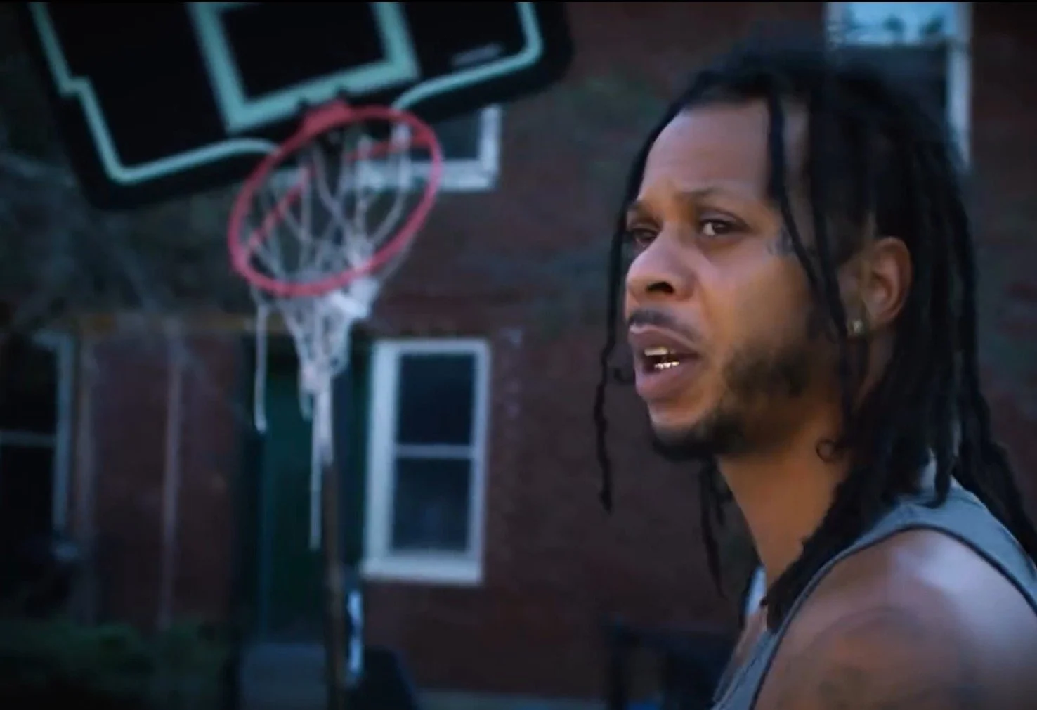 A man with long dreadlocks and earrings stands outdoors near a basketball hoop attached to a brick wall, looking to the side with a serious expression.