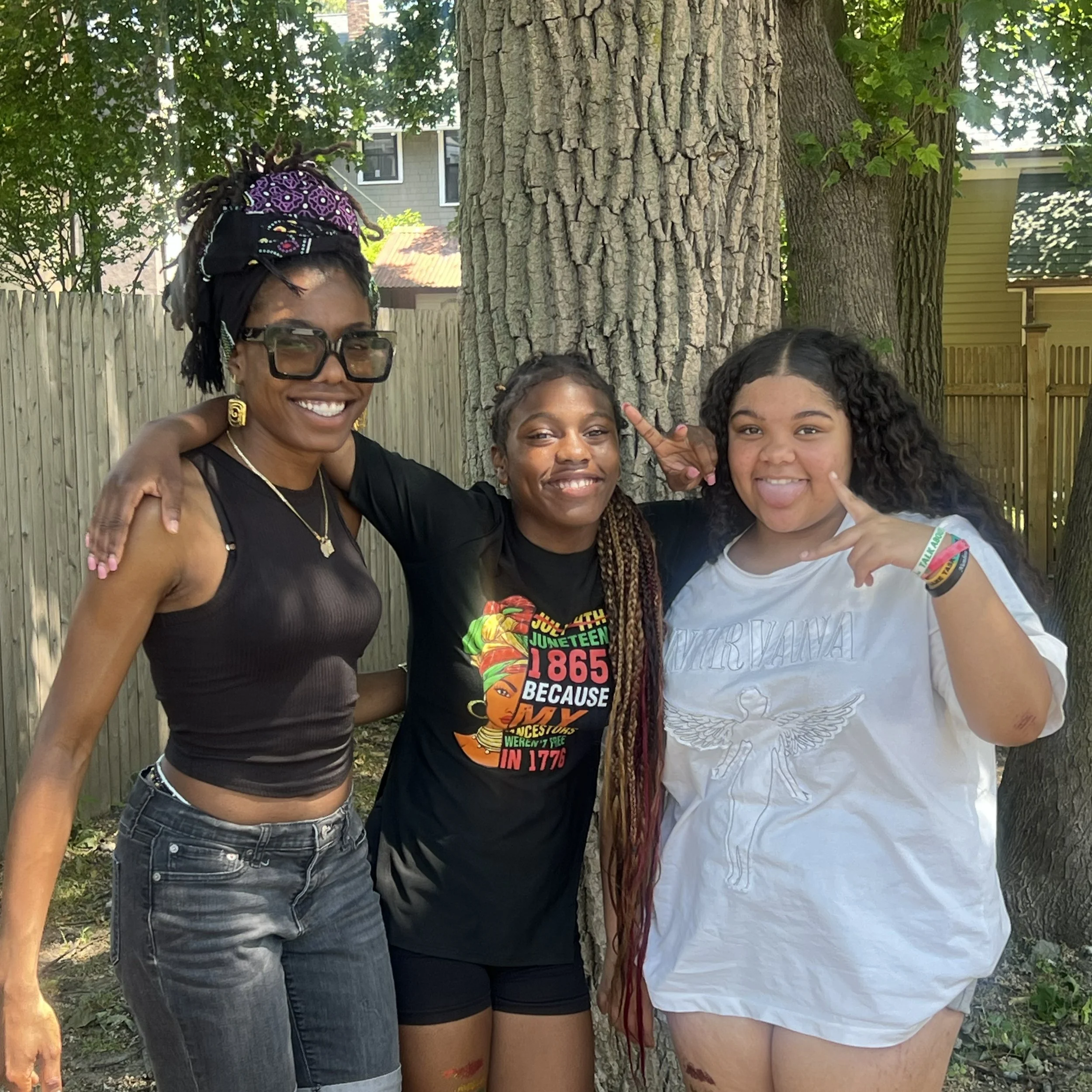 Four women standing together outdoors in front of a tree and a wooden fence, smiling at the camera.